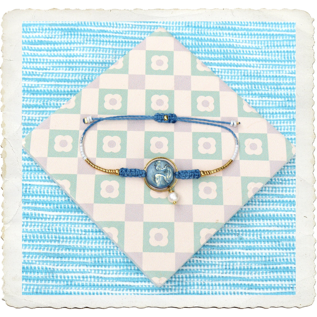 Peut inclure: Un bracelet bleu et or avec un charme de perle blanche et une perle de verre bleu et blanc. Le bracelet est sur un fond à carreaux blancs et verts.
