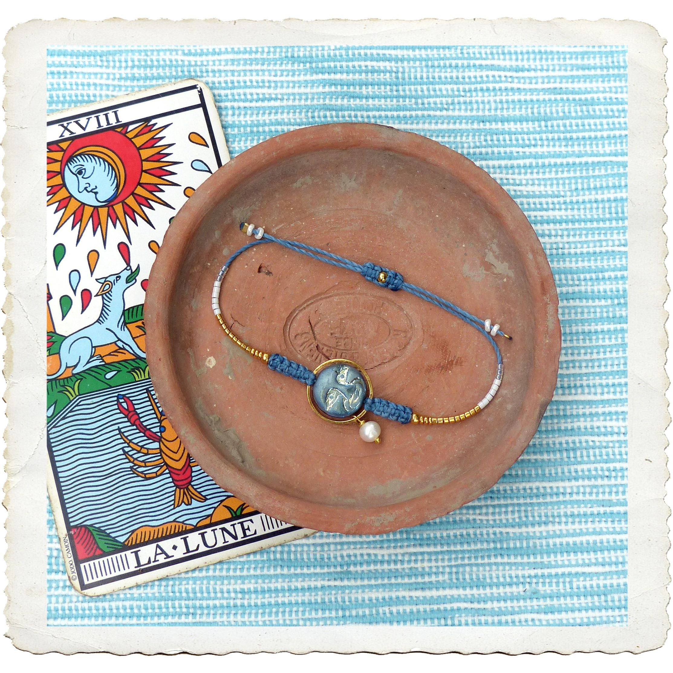 Peut inclure: Un bracelet bleu et or avec un charme camée bleu et blanc représentant une lune et des étoiles. Le bracelet est sur une assiette en terre cuite avec une carte de tarot représentant la lune en arrière-plan. La carte indique "XVIII LA LUNE".