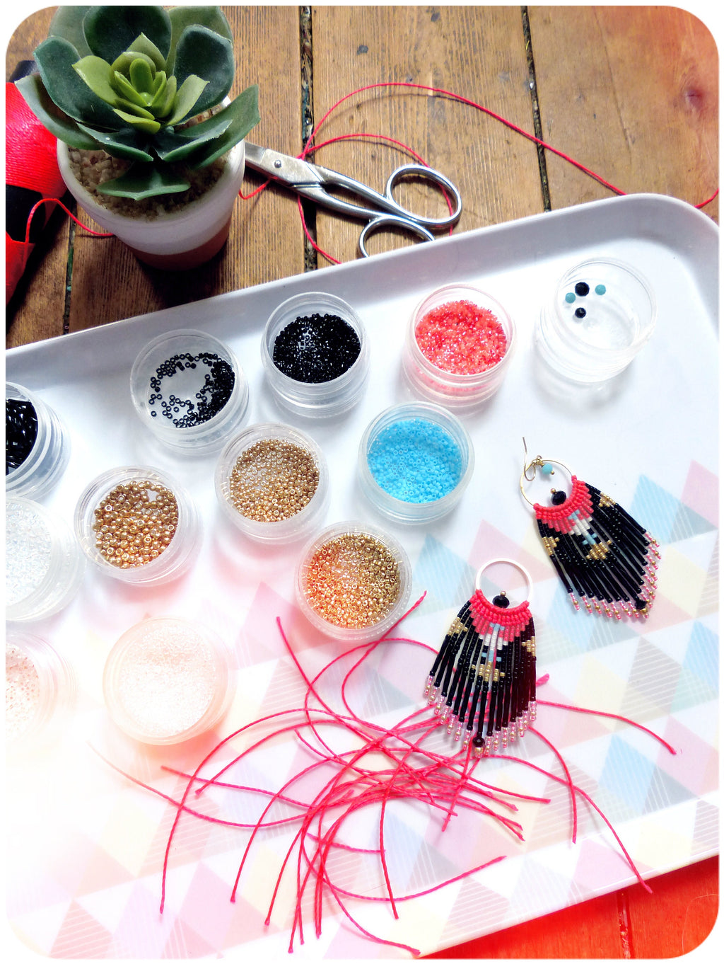 Peut inclure: Un plateau contenant de petits récipients remplis de perles colorées, ainsi que deux boucles d'oreilles en perles, une paire de ciseaux et une petite plante succulente en pot. Les boucles d'oreilles sont noires, blanches et roses avec un motif géométrique.
