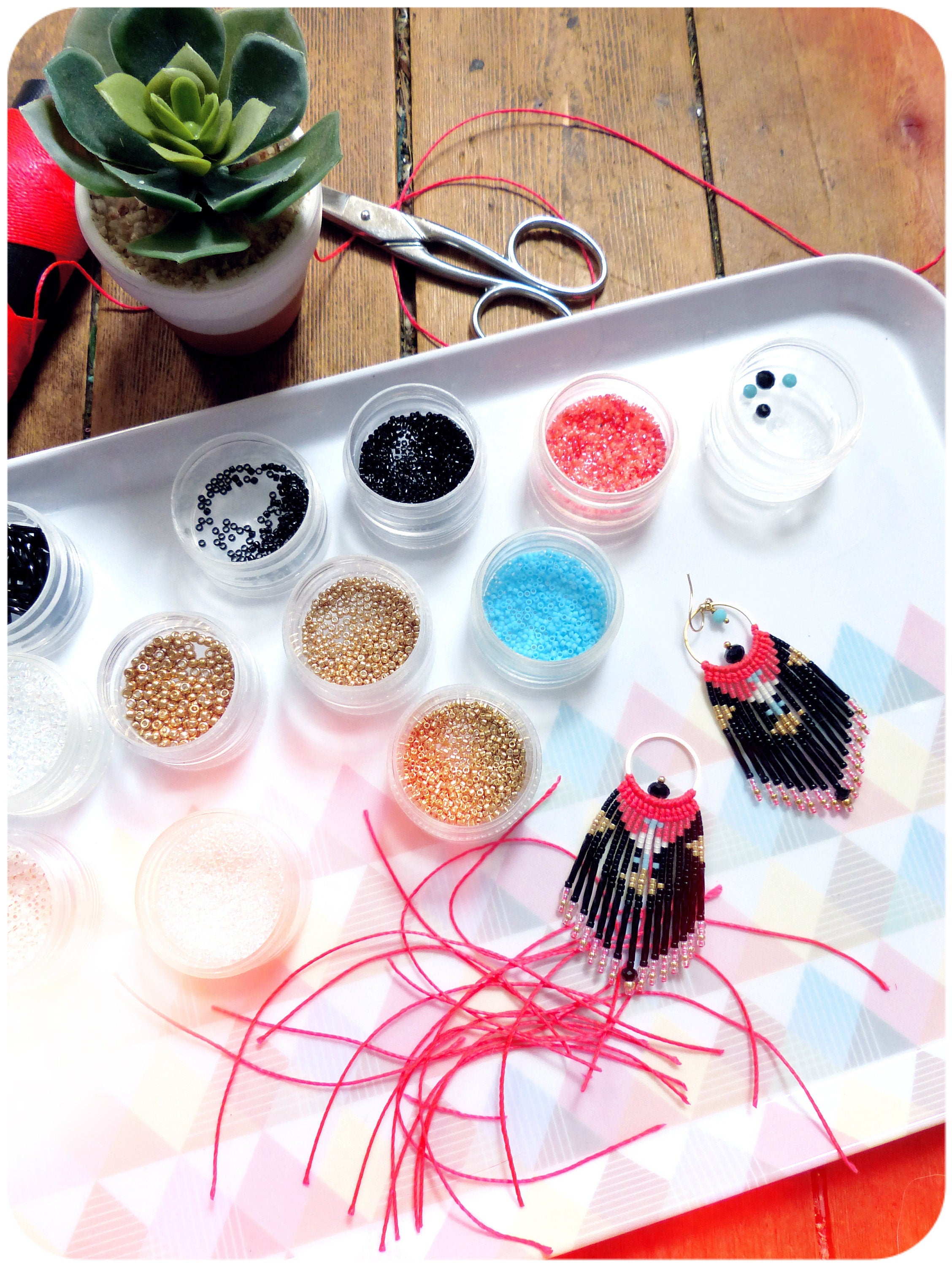 Peut inclure: Un plateau contenant de petits récipients remplis de perles colorées, ainsi que deux boucles d'oreilles en perles, une paire de ciseaux et une petite plante succulente en pot. Les boucles d'oreilles sont noires, blanches et roses avec un motif géométrique.