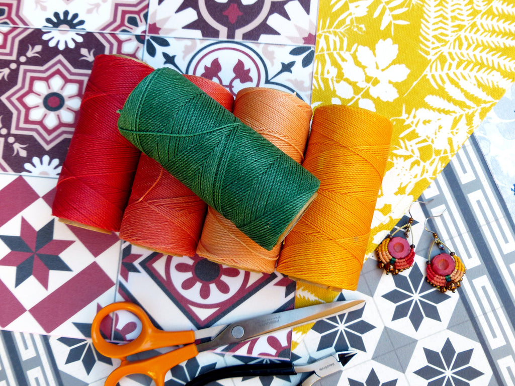 Peut inclure: Quatre bobines de fil coloré en rouge, orange, vert et jaune, avec une paire de boucles d'oreilles et une paire de ciseaux sur un fond de carrelage à motifs.