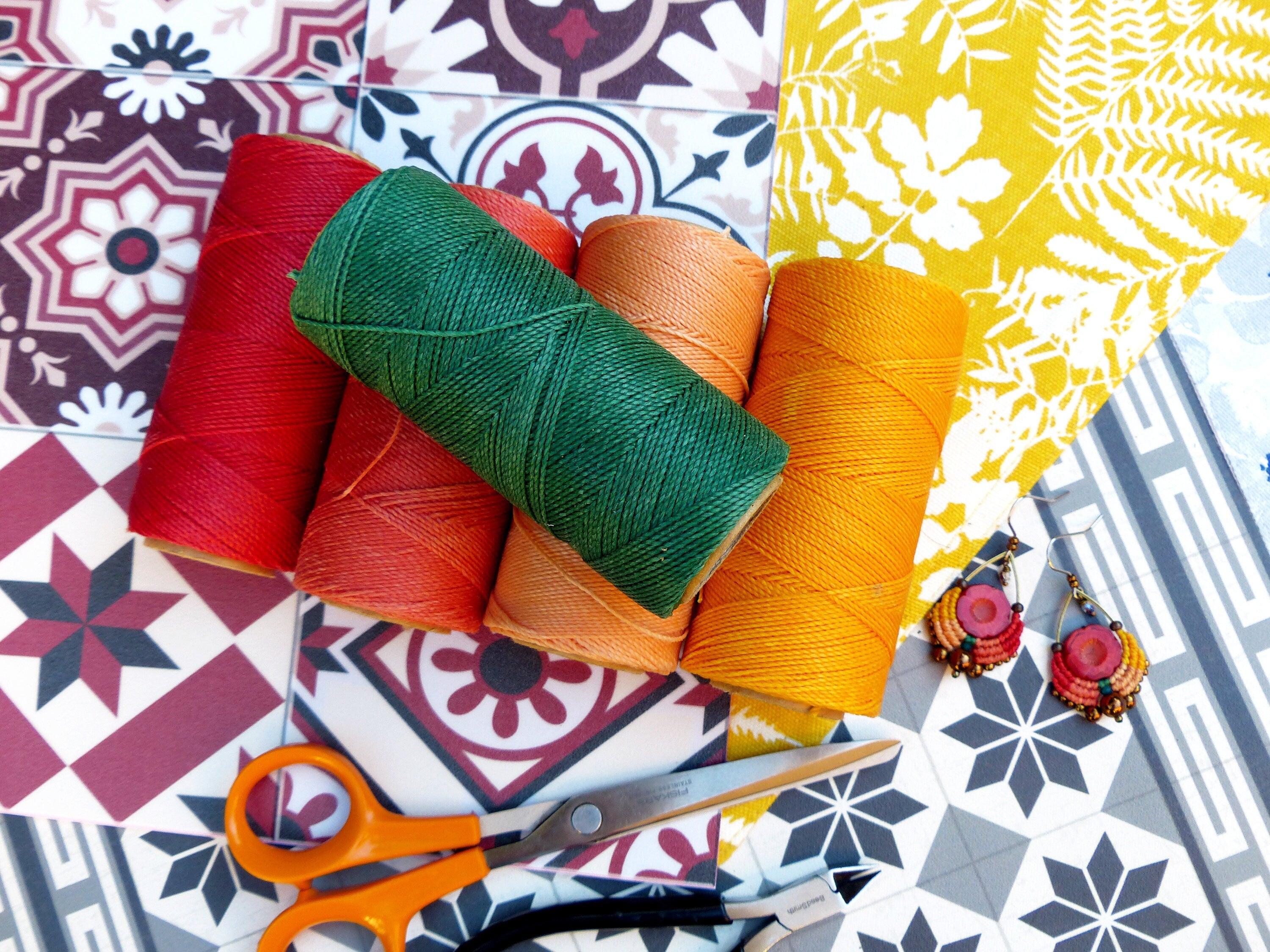 Peut inclure: Quatre bobines de fil coloré en rouge, orange, vert et jaune, avec une paire de boucles d'oreilles et une paire de ciseaux sur un fond de carrelage à motifs.
