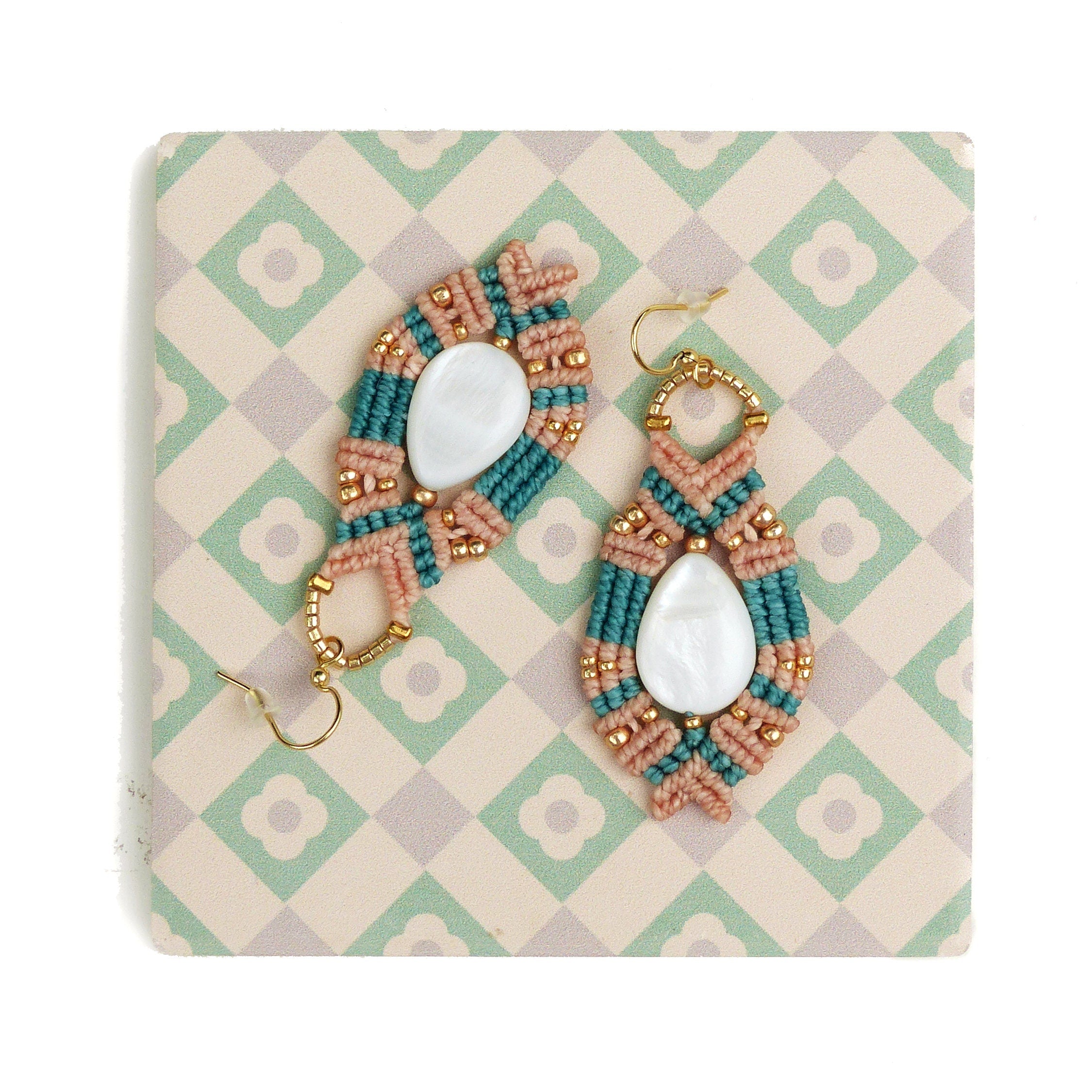 Peut inclure: Une paire de boucles d'oreilles faites à la main avec une perle de coquille blanche en forme de larme au centre. Les boucles d'oreilles sont réalisées avec un cordon de macramé rose et turquoise et des crochets en métal doré.