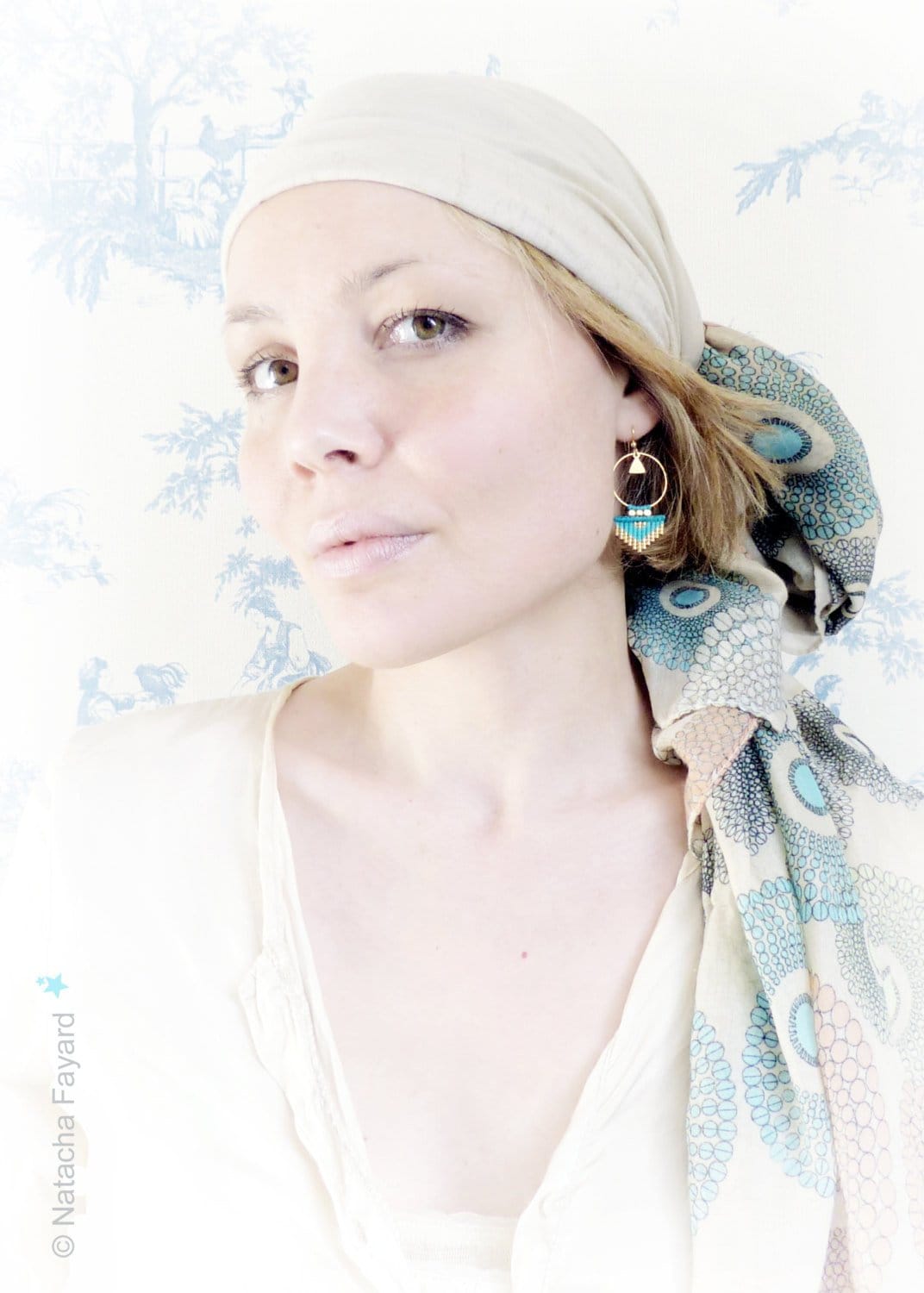 Peut inclure: Une femme portant un foulard blanc sur la tête et un foulard à motifs noué autour du cou. Elle porte une paire de boucles d'oreilles pendantes dorées et bleues.