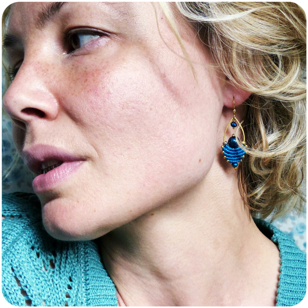 Peut inclure: Une femme portant des boucles d'oreilles bleues avec des accents dorés et des perles bleues. Les boucles d'oreilles sont pendantes et ont une forme de larme.