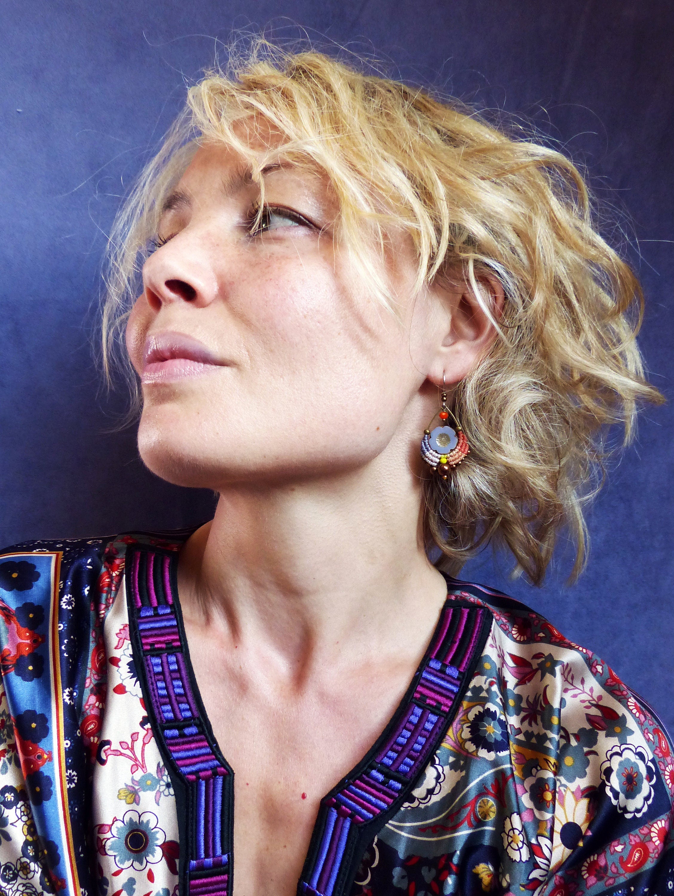 Peut inclure: Une femme aux cheveux blonds portant un chemisier à motifs colorés avec un col brodé de violet et de bleu. Elle porte une paire de boucles d'oreilles pendantes avec des perles bleues, orange et rouges.