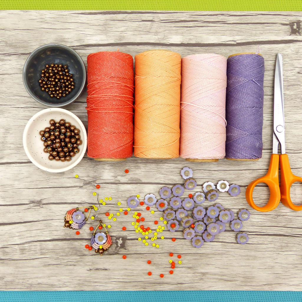 Peut inclure: Une vue de dessus de fournitures artisanales pour la fabrication de bijoux. Quatre bobines de fil dans des tons d'orange, rose et violet. Deux petits bols de perles brunes. Une paire de boucles d'oreilles avec des perles violettes et orange. Une dispersion de perles orange et jaunes. Une paire de ciseaux.