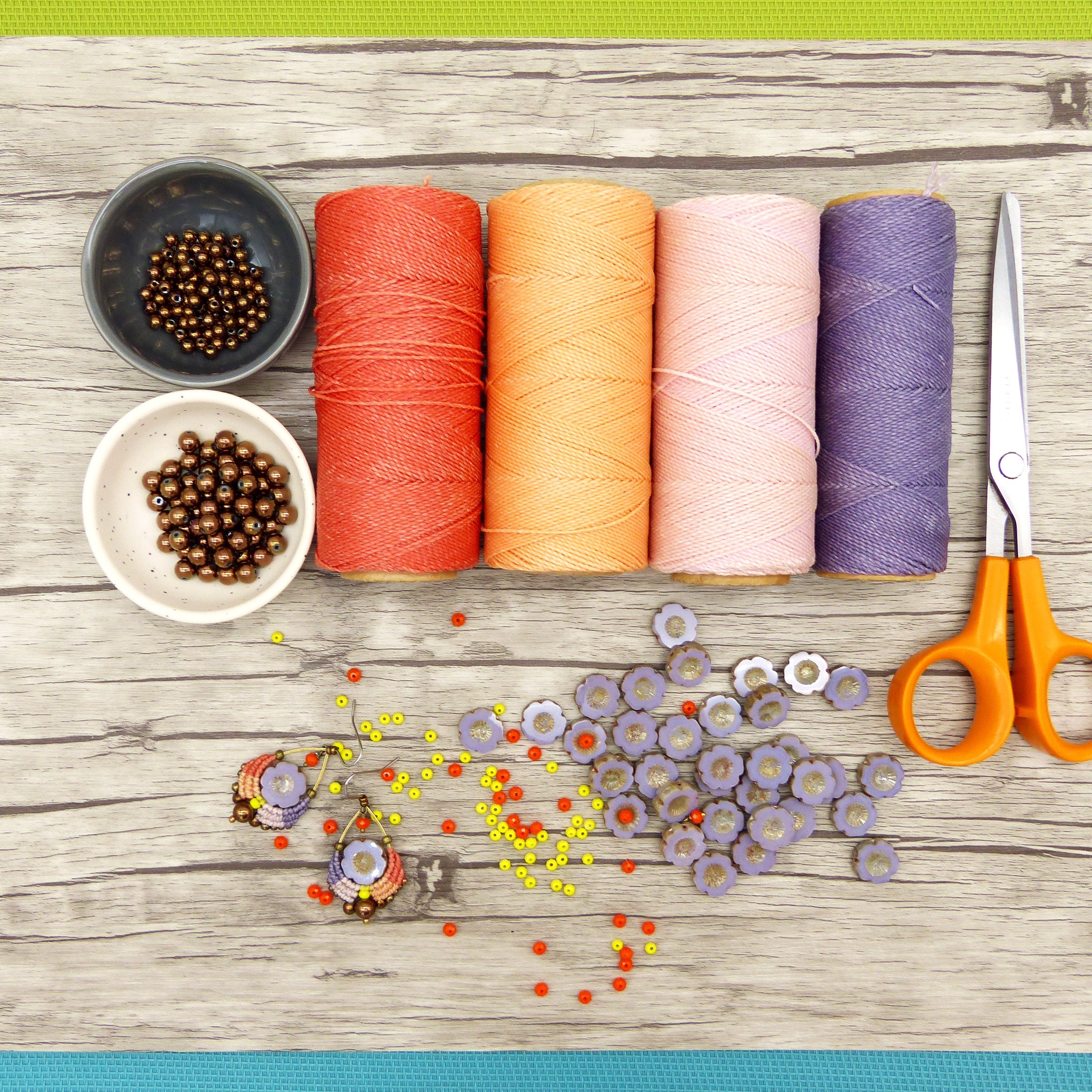 Peut inclure: Une vue de dessus de fournitures artisanales pour la fabrication de bijoux. Quatre bobines de fil dans des tons d'orange, rose et violet. Deux petits bols de perles brunes. Une paire de boucles d'oreilles avec des perles violettes et orange. Une dispersion de perles orange et jaunes. Une paire de ciseaux.