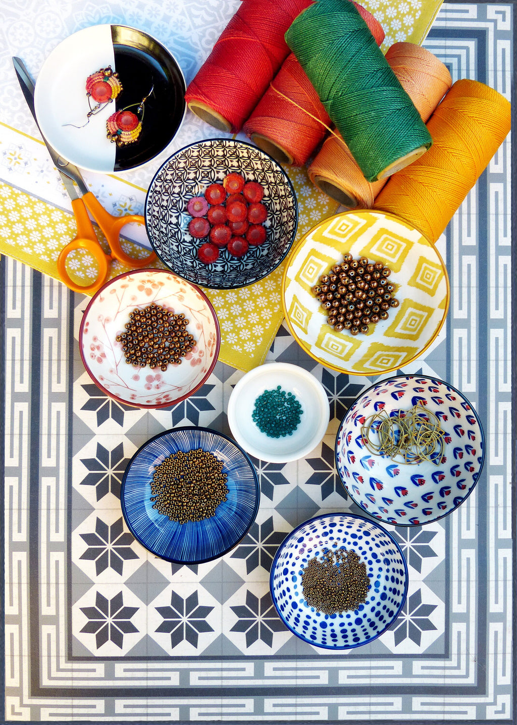 Peut inclure: Assortiment de bols colorés remplis de perles et d'autres fournitures d'artisanat. Les bols sont disposés sur une surface carrelée avec un motif floral. Il y a aussi des bobines de fil de différentes couleurs.