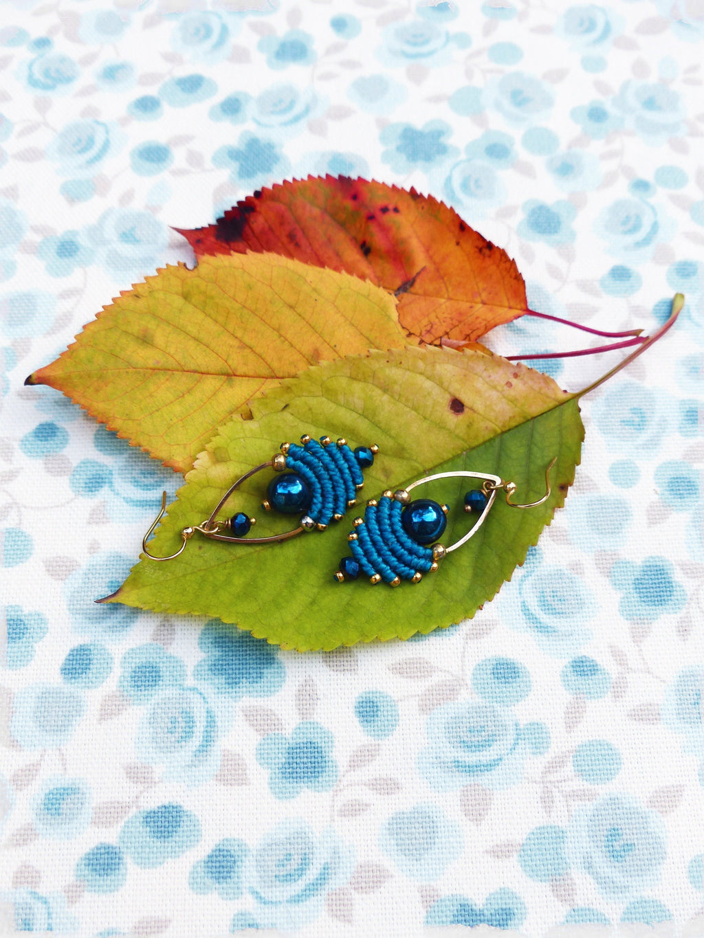 Peut inclure: Une paire de boucles d'oreilles bleues et dorées avec un motif perlé. Les boucles d'oreilles sont sur un fond blanc avec un motif floral.