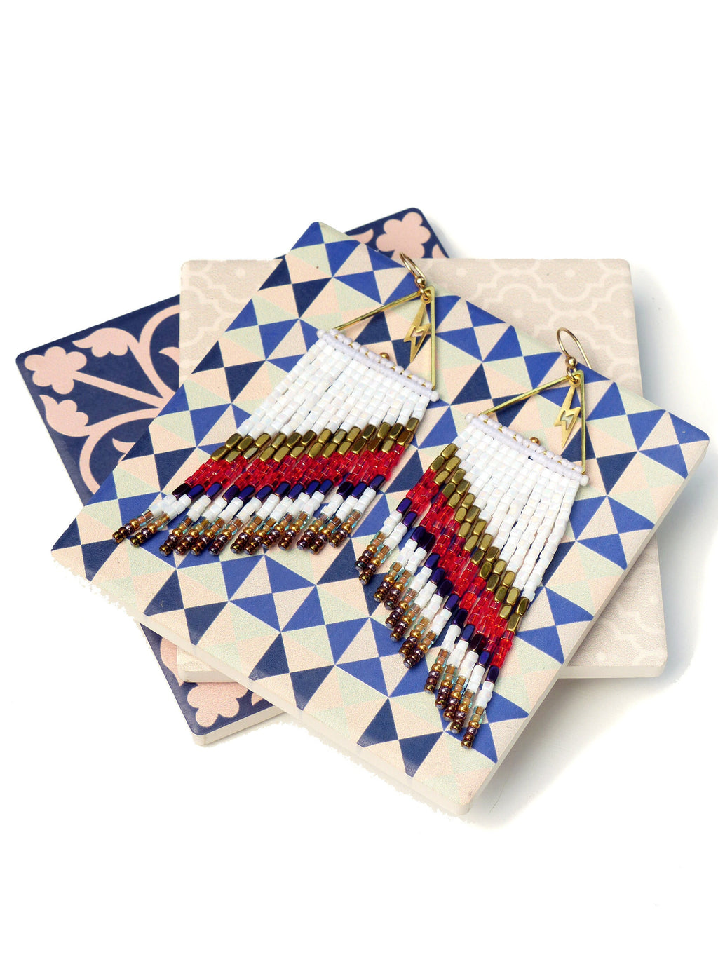 Peut inclure: Une paire de longues boucles d'oreilles en perles blanches avec des accents rouges, bleus et dorés. Les boucles d'oreilles sont en forme de triangle et ont une frange de perles.