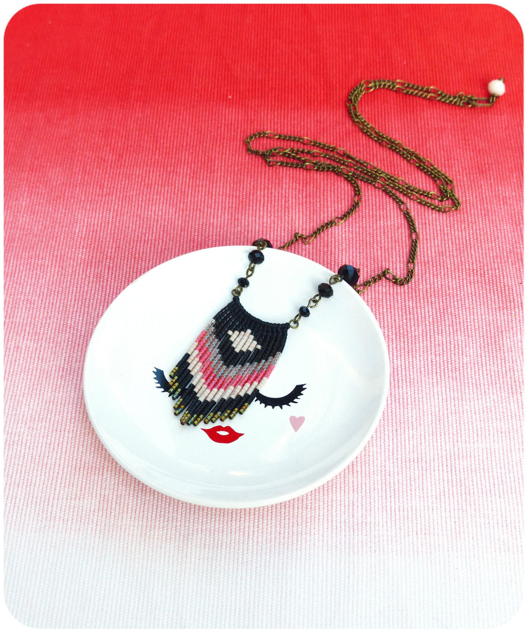 Peut inclure: Un collier de perles noires, roses et blanches avec un pendentif en forme de frange est suspendu à un plat en céramique blanche avec un motif de visage stylisé. Le collier est sur une chaîne dorée.