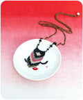 Peut inclure: Un collier de perles noires, roses et blanches avec un pendentif en forme de frange est suspendu à un plat en céramique blanche avec un motif de visage stylisé. Le collier est sur une chaîne dorée.