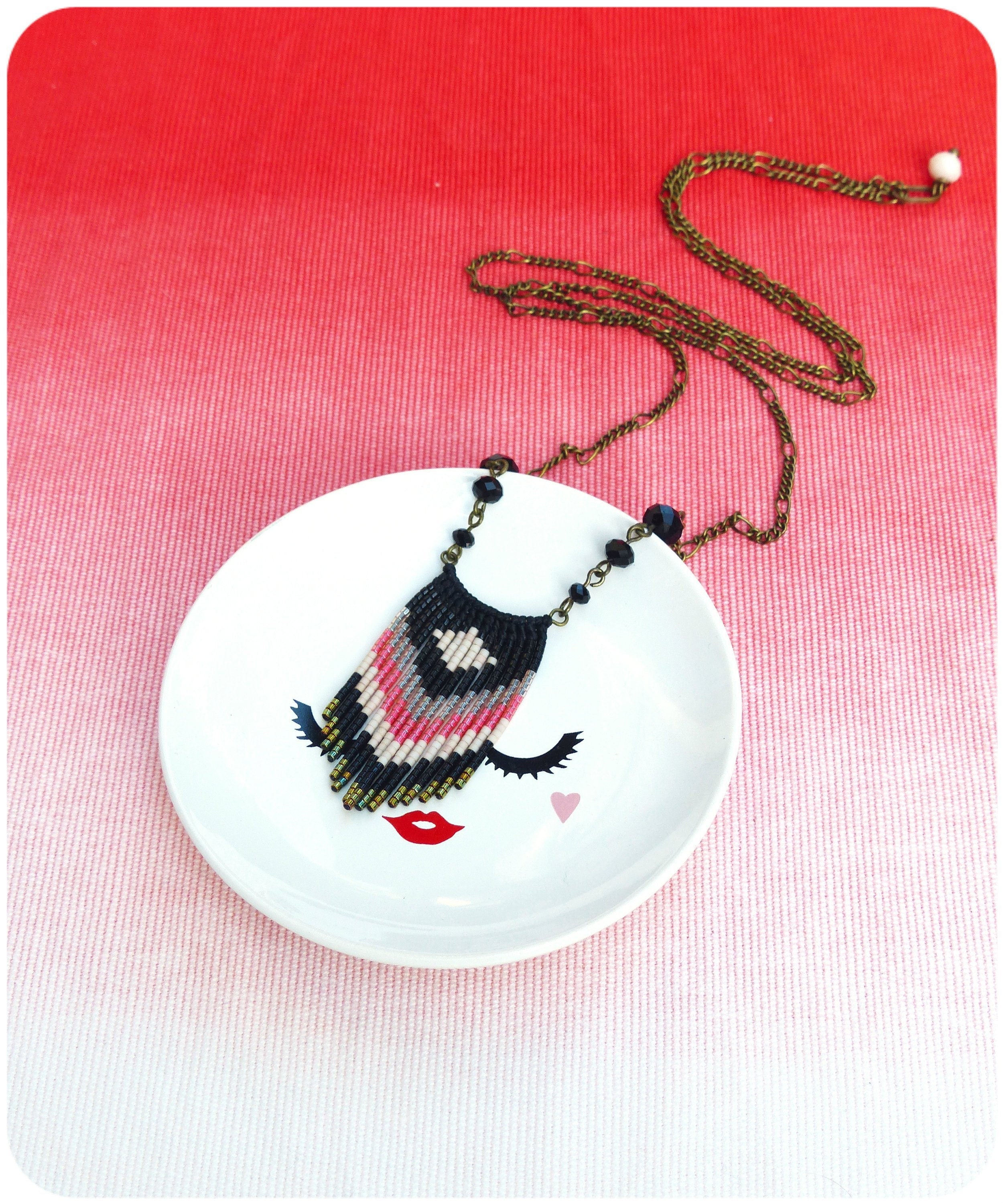 Peut inclure: Un collier de perles noires, roses et blanches avec un pendentif en forme de frange est suspendu à un plat en céramique blanche avec un motif de visage stylisé. Le collier est sur une chaîne dorée.