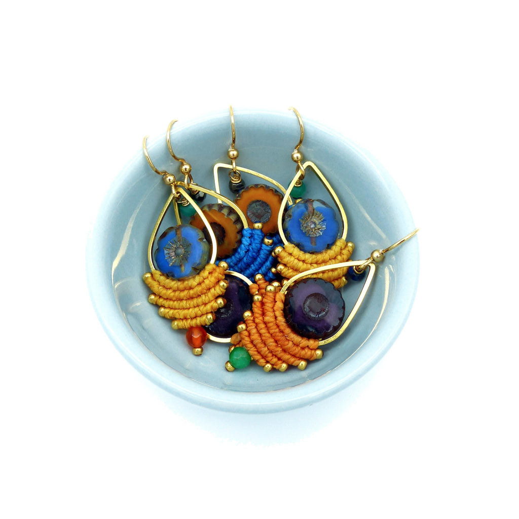 Peut inclure: Un ensemble de cinq boucles d'oreilles colorées avec des anneaux dorés et une variété de perles bleues, violettes, orange et jaunes. Les boucles d'oreilles sont présentées dans un petit bol bleu clair.