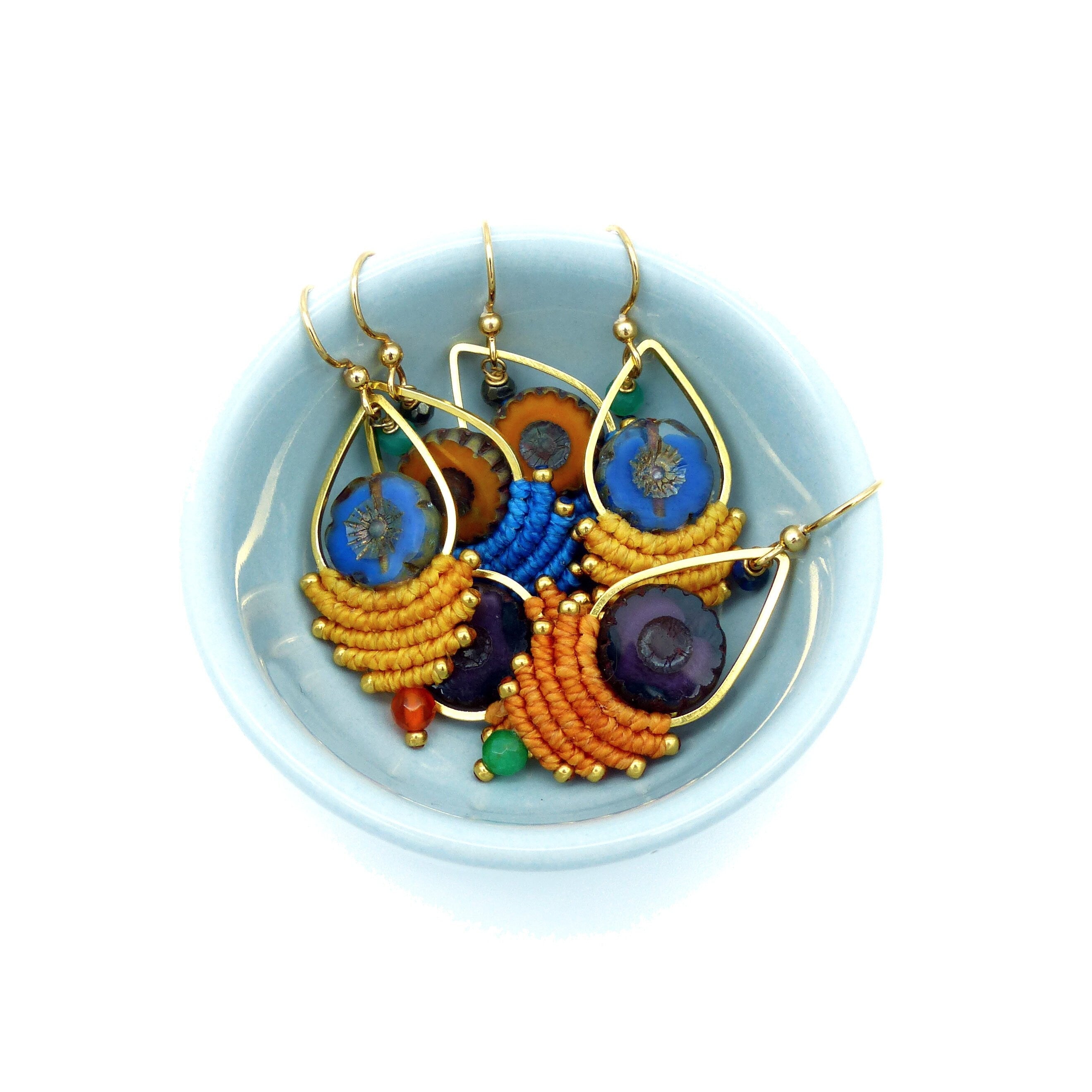 Peut inclure: Un ensemble de cinq boucles d'oreilles colorées avec des anneaux dorés et une variété de perles bleues, violettes, orange et jaunes. Les boucles d'oreilles sont présentées dans un petit bol bleu clair.