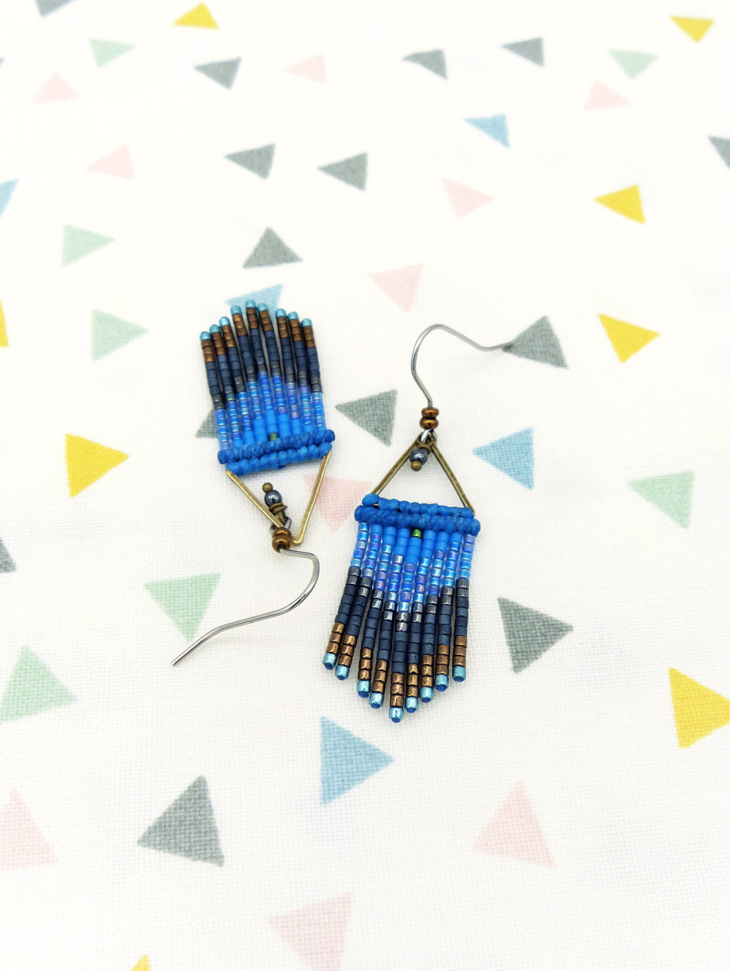 Peut inclure: Une paire de boucles d'oreilles en perles bleues et dorées avec un design triangulaire. Les boucles d'oreilles sont suspendues à des crochets argentés.