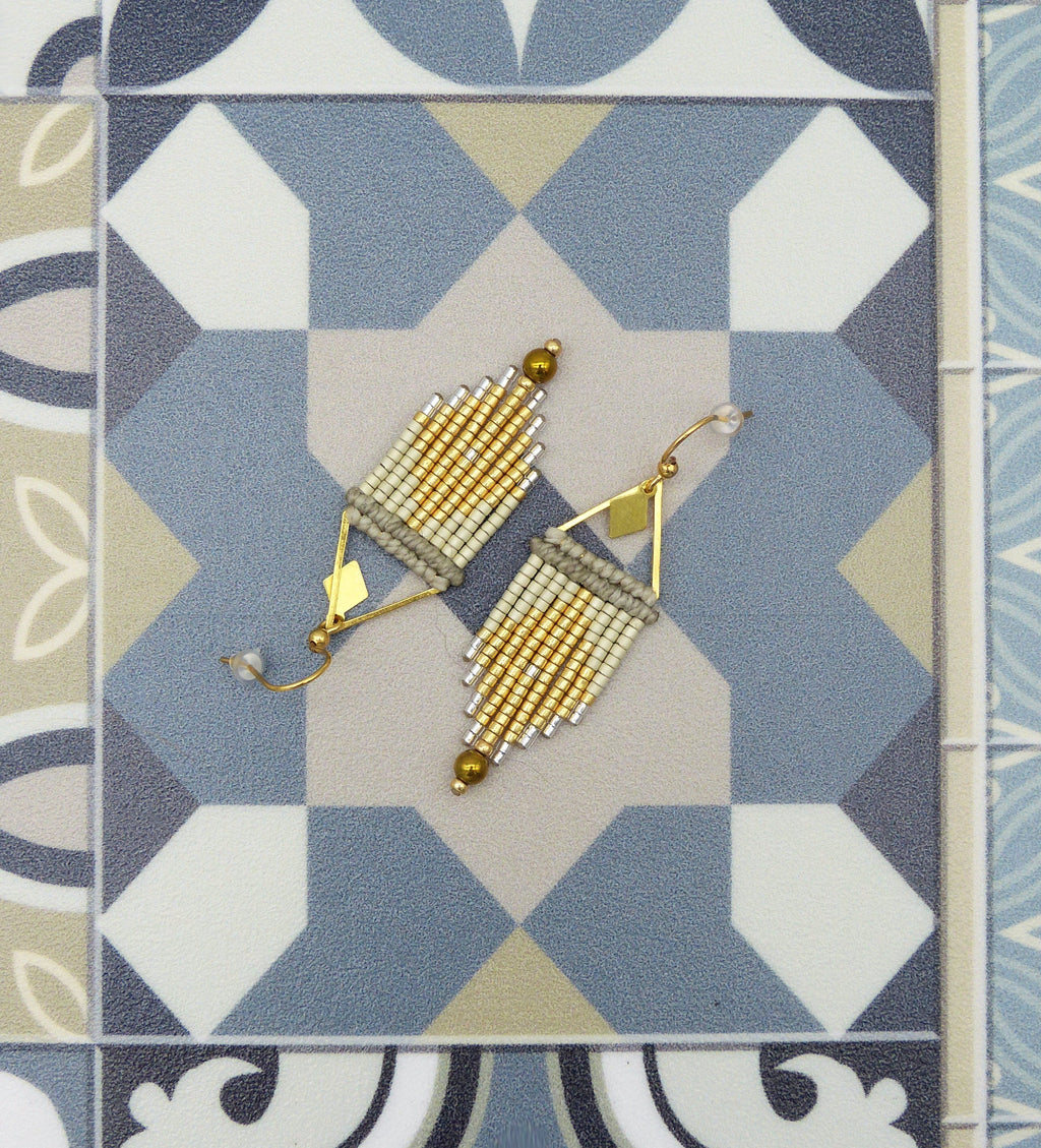 Peut inclure: Une paire de boucles d'oreilles en perles dorées et blanches avec un motif géométrique. Les boucles d'oreilles sont suspendues à une surface carrelée bleue et blanche.