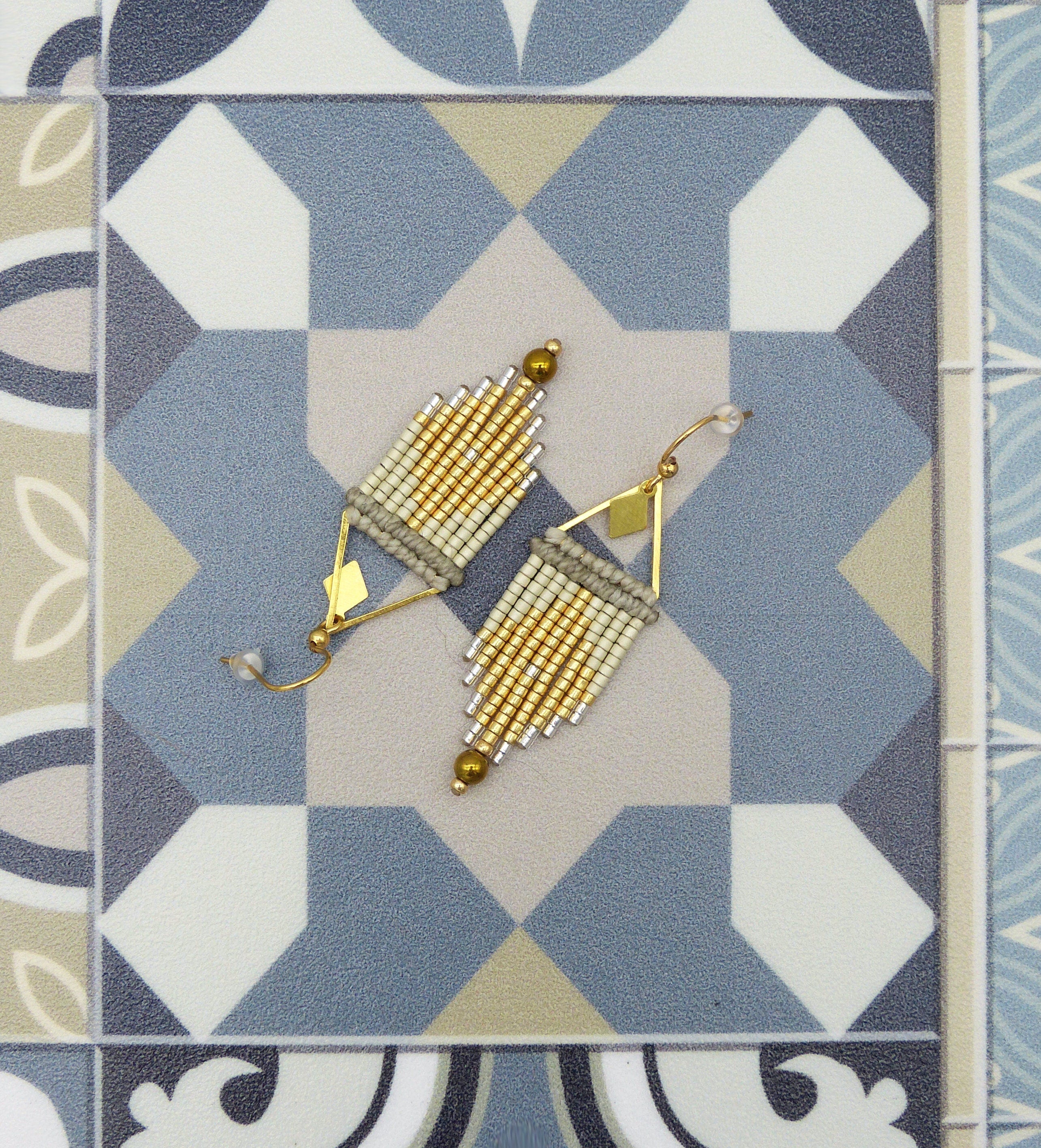 Peut inclure: Une paire de boucles d'oreilles en perles dorées et blanches avec un motif géométrique. Les boucles d'oreilles sont suspendues à une surface carrelée bleue et blanche.