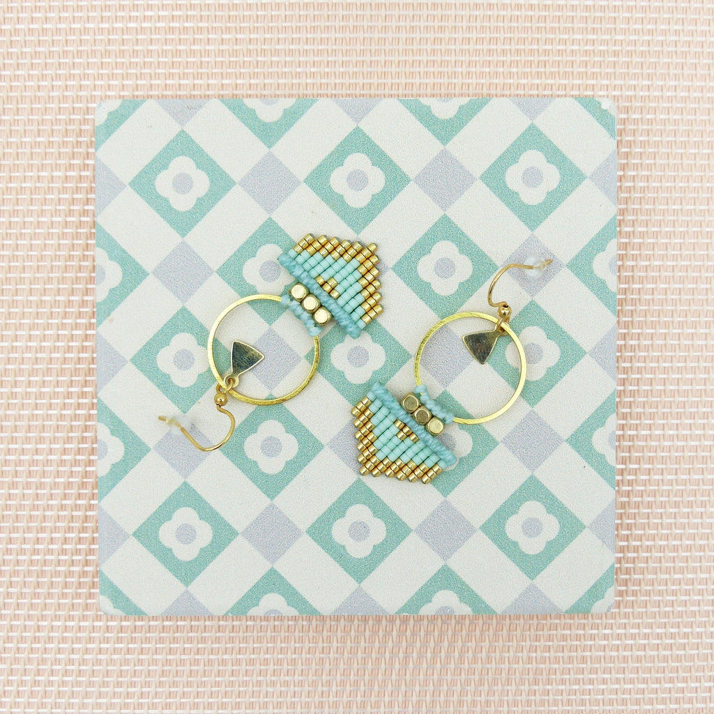 Peut inclure: Une paire de boucles d'oreilles créoles dorées avec des formes géométriques en perles bleues et dorées qui en pendent. Les boucles d'oreilles sont présentées sur une surface carrelée blanche et verte.