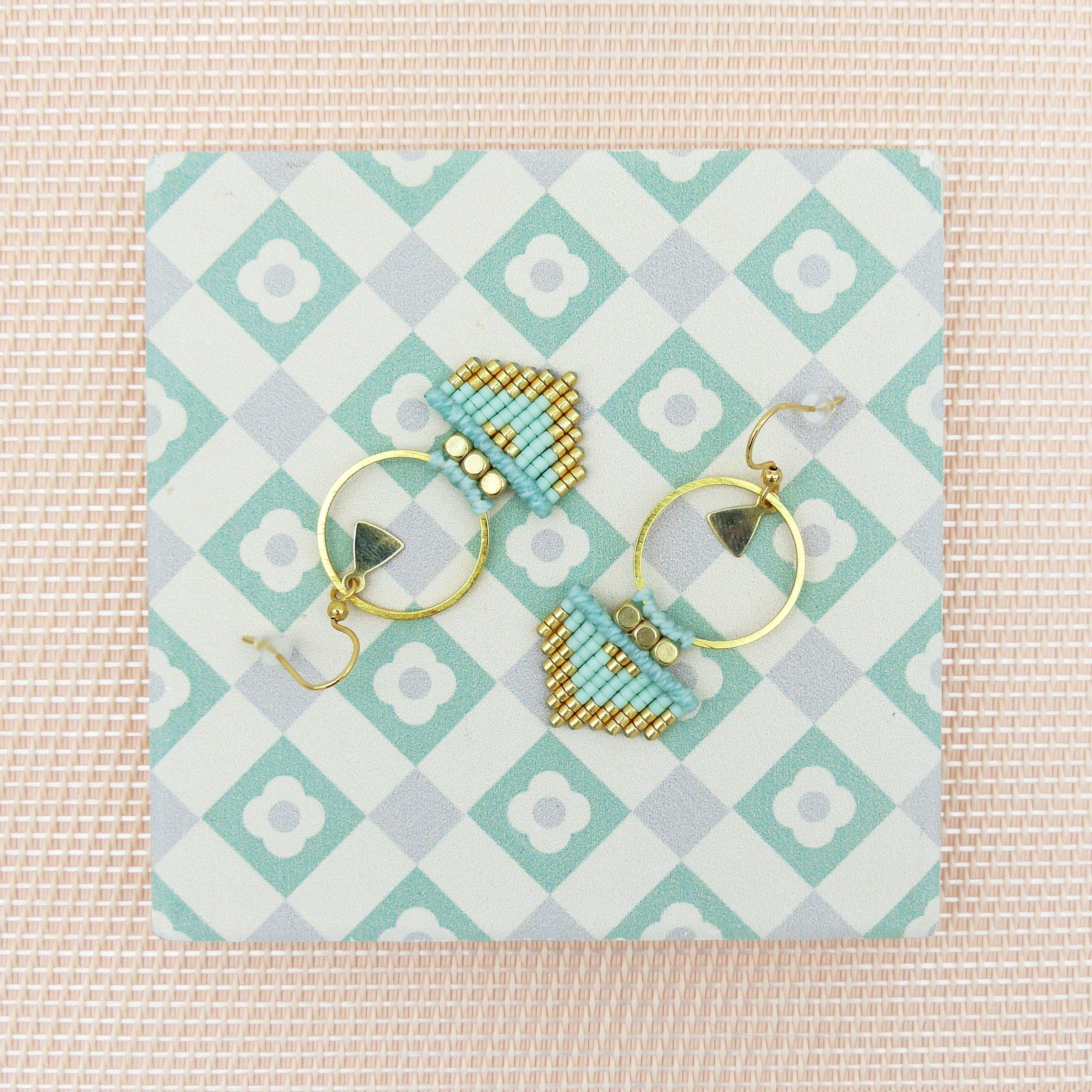 Peut inclure: Une paire de boucles d'oreilles créoles dorées avec des formes géométriques en perles bleues et dorées qui en pendent. Les boucles d'oreilles sont présentées sur une surface carrelée blanche et verte.
