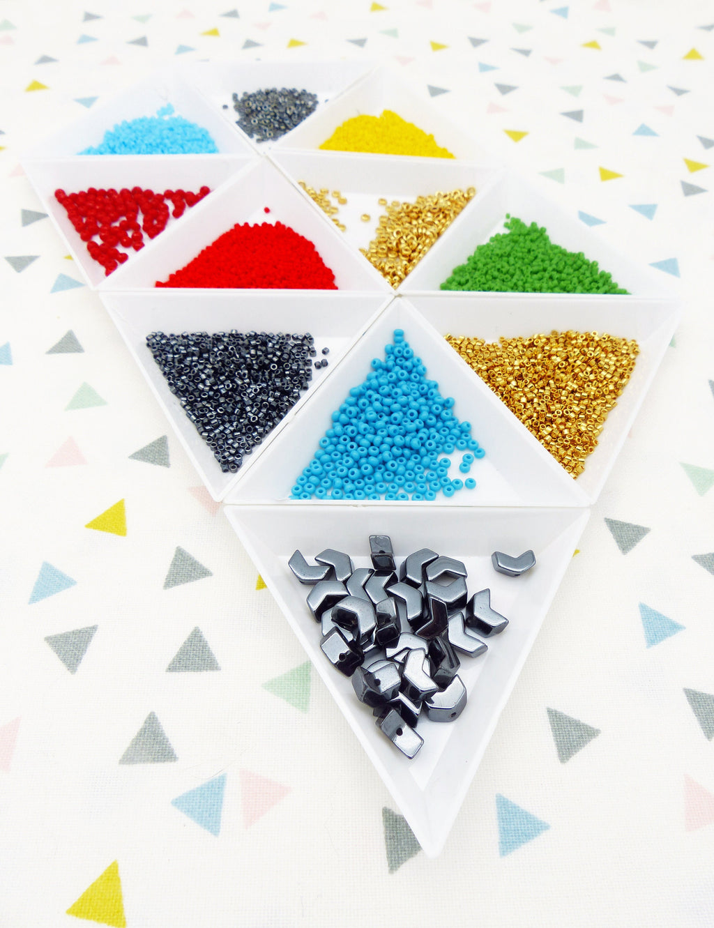 Peut inclure: Un ensemble de neuf plateaux triangulaires blancs remplis de perles colorées. Les plateaux sont disposés en cercle avec un plus petit plateau au centre contenant des perles hexagonales noires.