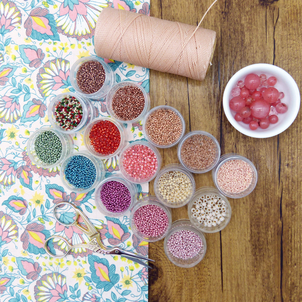 Peut inclure: Assortiment de petits contenants remplis de perles colorées, disposés sur une surface en bois. Une bobine de fil et un bol de perles roses sont également visibles.