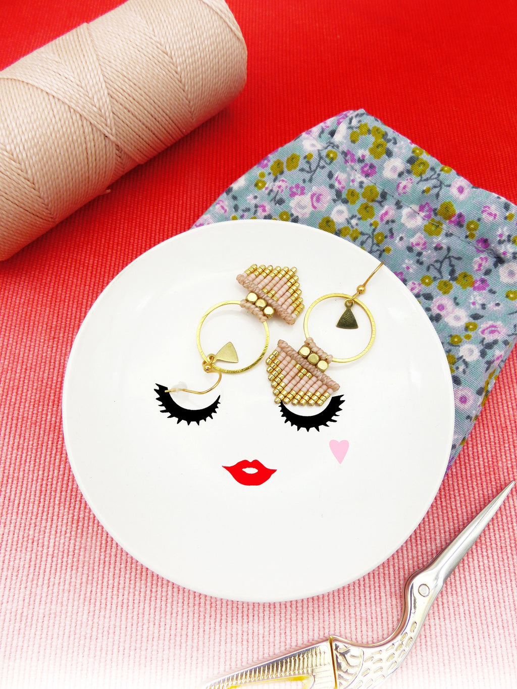Peut inclure: Un plat en céramique blanche avec un design de visage stylisé comprenant des cils, des lèvres et un cœur rose. Deux paires de boucles d'oreilles en perles dorées et roses sont exposées sur le plat.