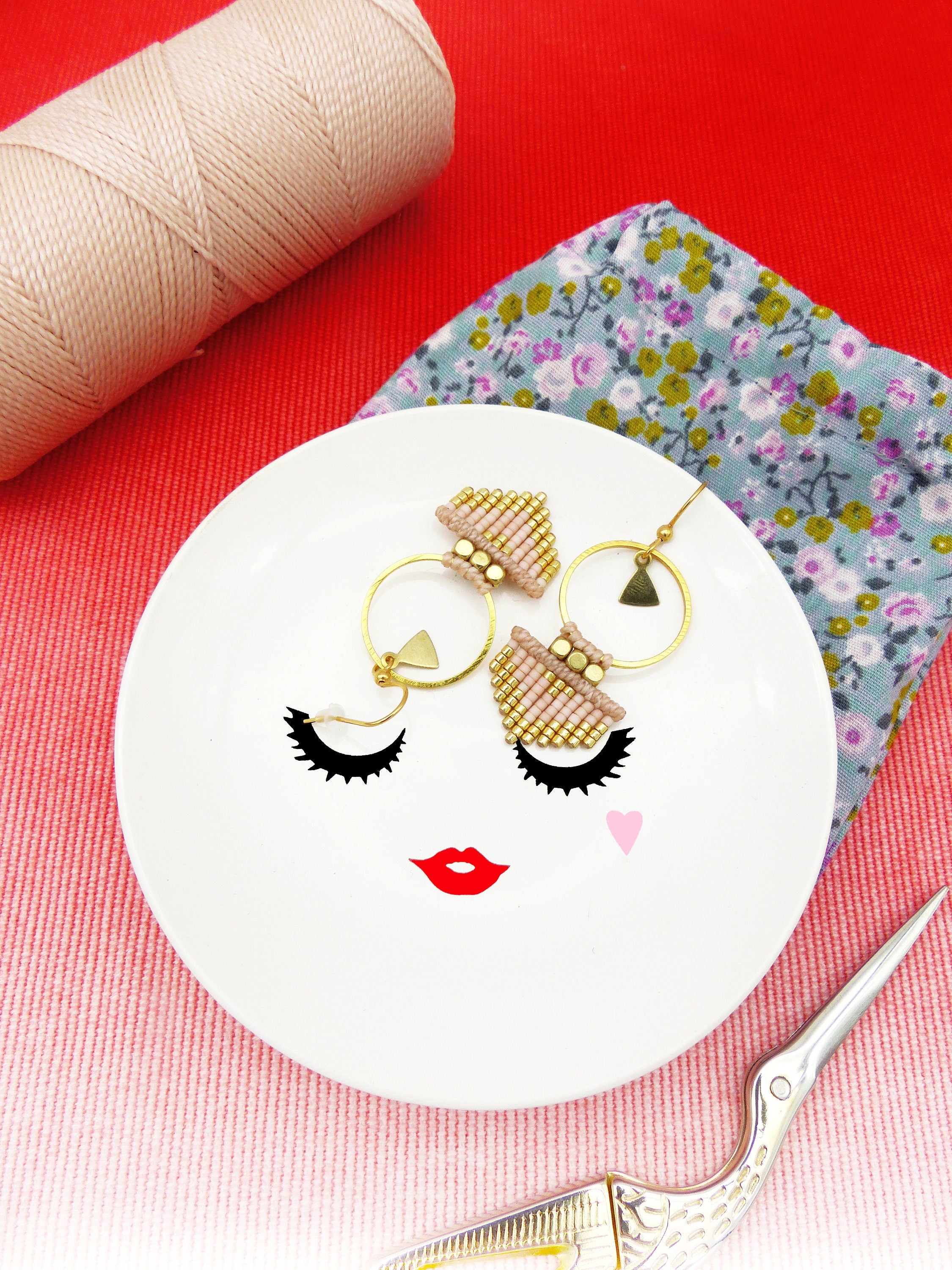 Peut inclure: Un plat en céramique blanche avec un design de visage stylisé comprenant des cils, des lèvres et un cœur rose. Deux paires de boucles d'oreilles en perles dorées et roses sont exposées sur le plat.