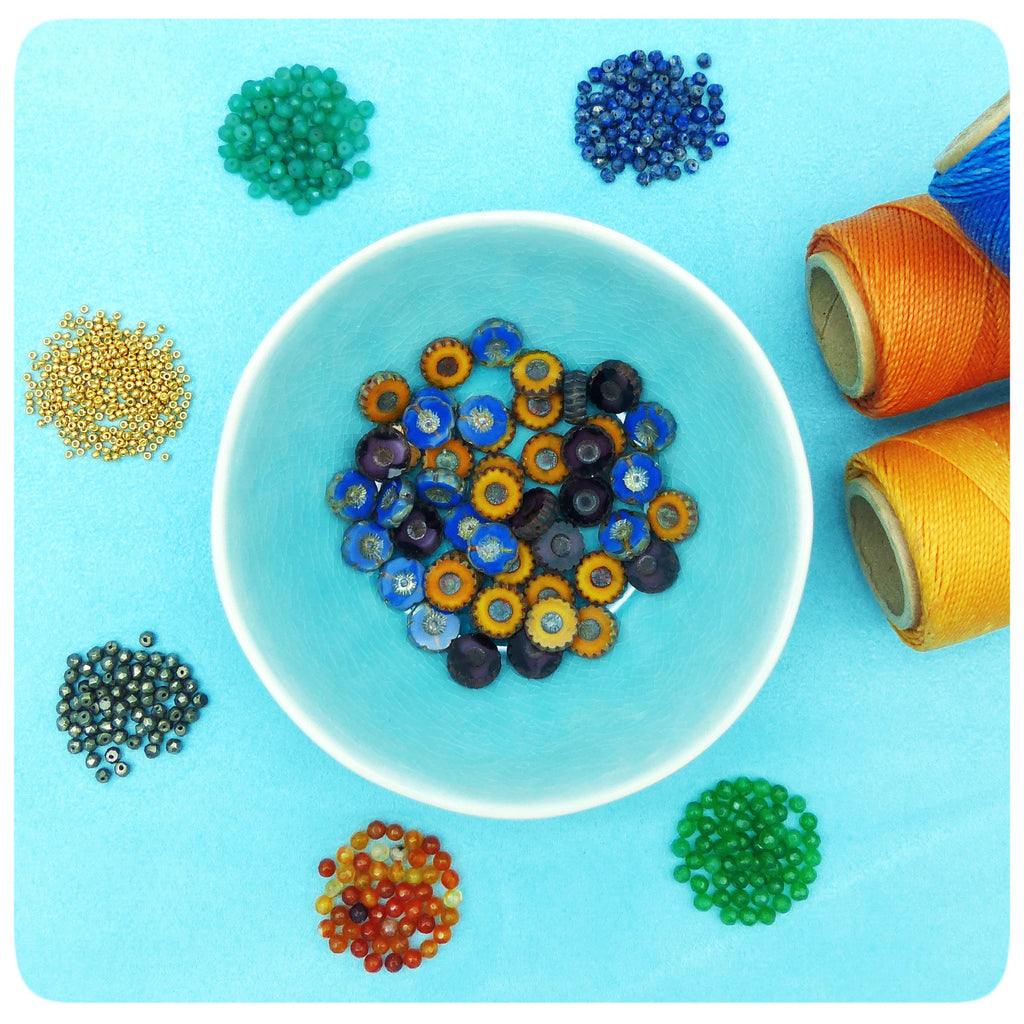 Peut inclure: Assortiment de perles de verre colorées de différentes formes et tailles, notamment des perles rondes, carrées et en forme d'étoile. Les perles sont disposées dans de petits bols sur une surface bleu clair. Il y a aussi deux bobines de fil, une orange et une jaune.