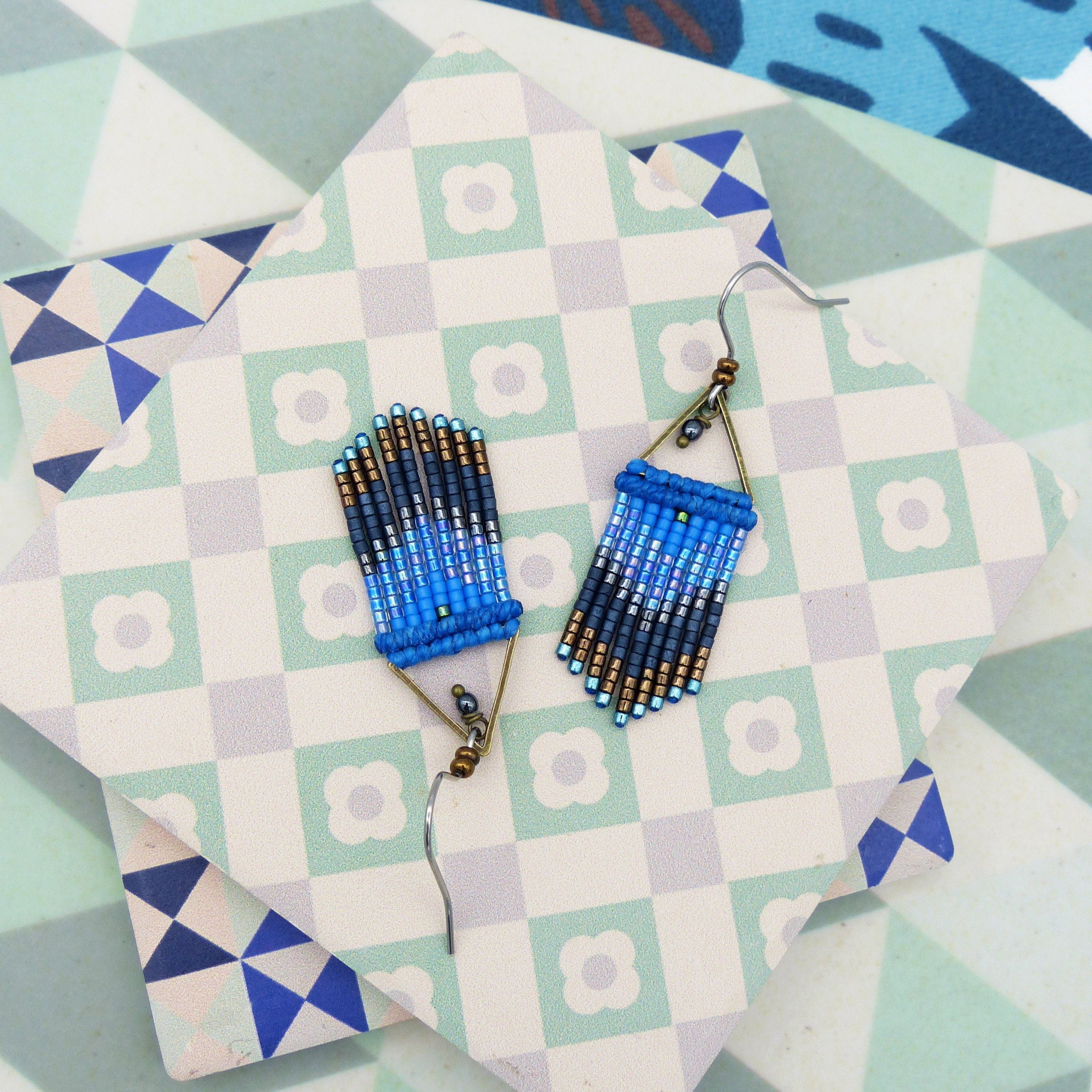Peut inclure: Une paire de boucles d'oreilles en franges de perles bleues et noires avec des crochets en métal doré. Les boucles d'oreilles sont suspendues à une surface carrelée blanche et verte.