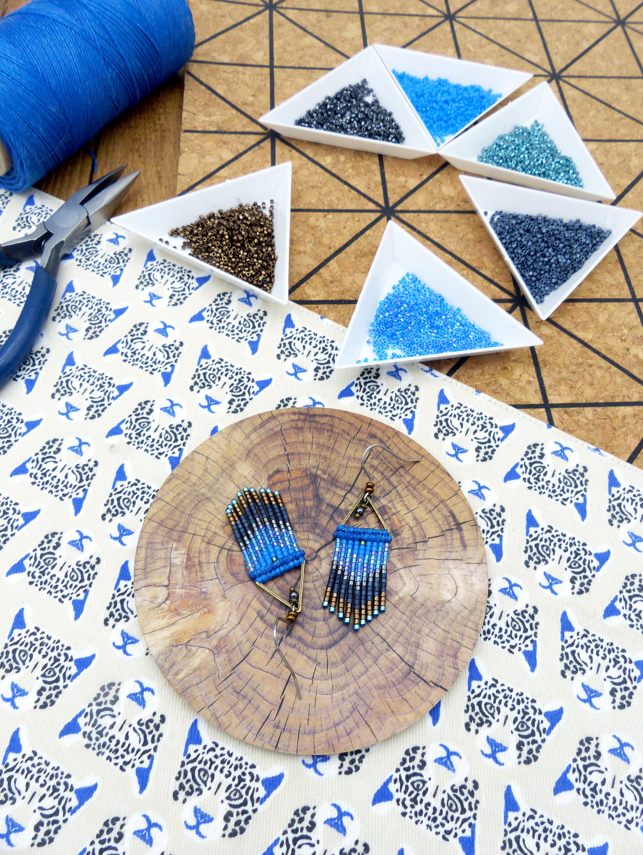Peut inclure: Une paire de boucles d'oreilles en perles bleues et brunes de forme triangulaire, disposées sur une surface en bois. Les boucles d'oreilles sont entourées de petits récipients de perles de différentes couleurs.