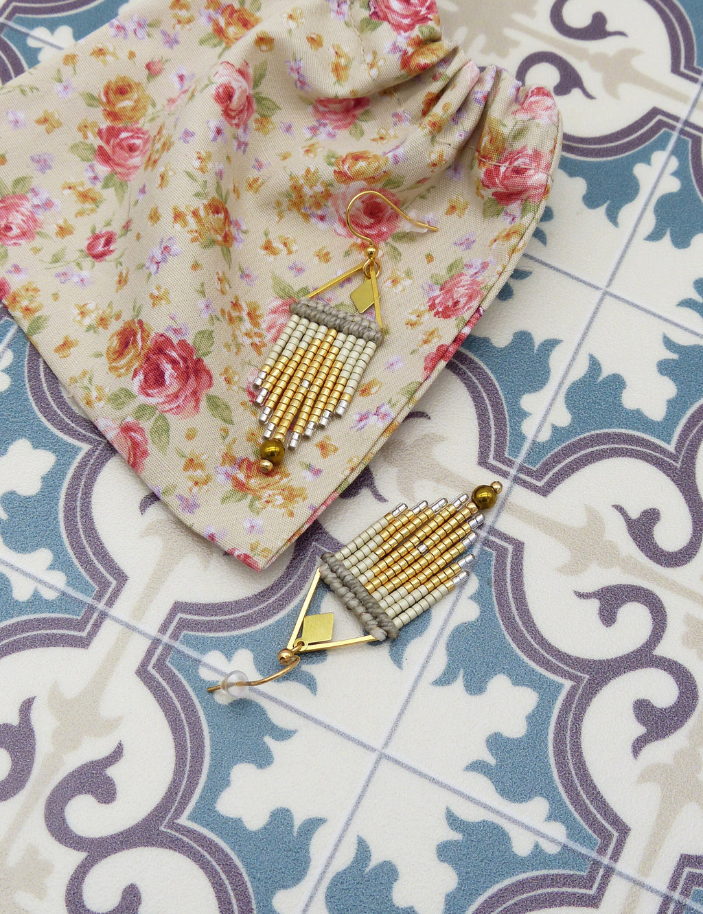 Peut inclure: Une paire de boucles d'oreilles en forme de triangle, composées de perles dorées et blanches. Les boucles d'oreilles sont suspendues à un sac en tissu fleuri.