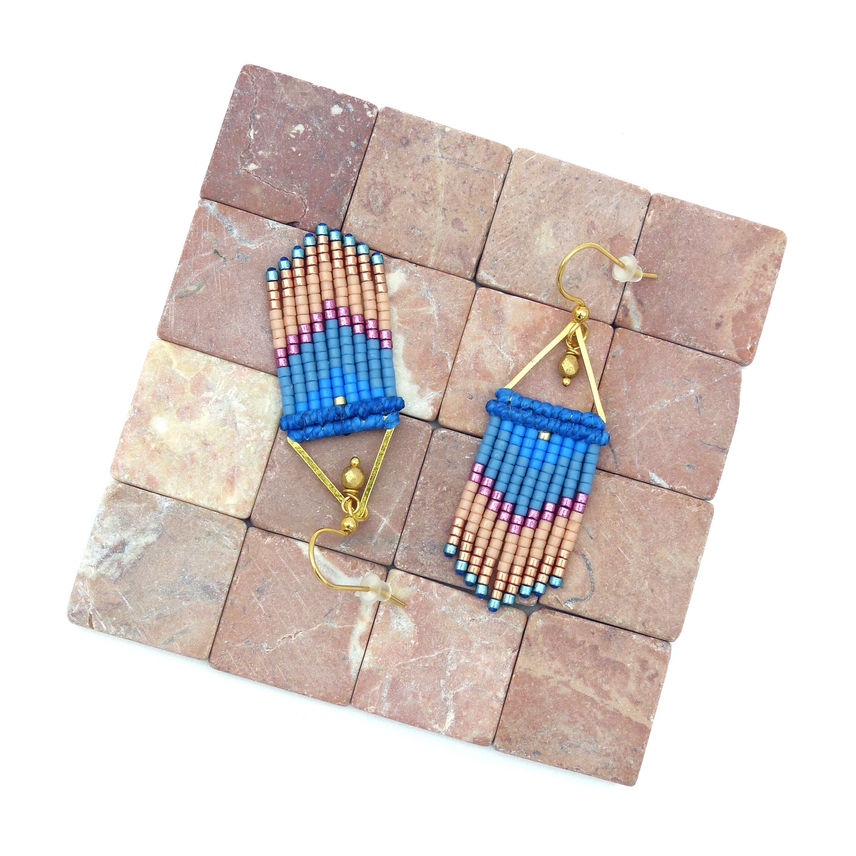 Peut inclure: Une paire de boucles d'oreilles dorées avec des franges de perles bleues, roses et pêche. Les boucles d'oreilles sont présentées sur une surface de carreaux bruns.