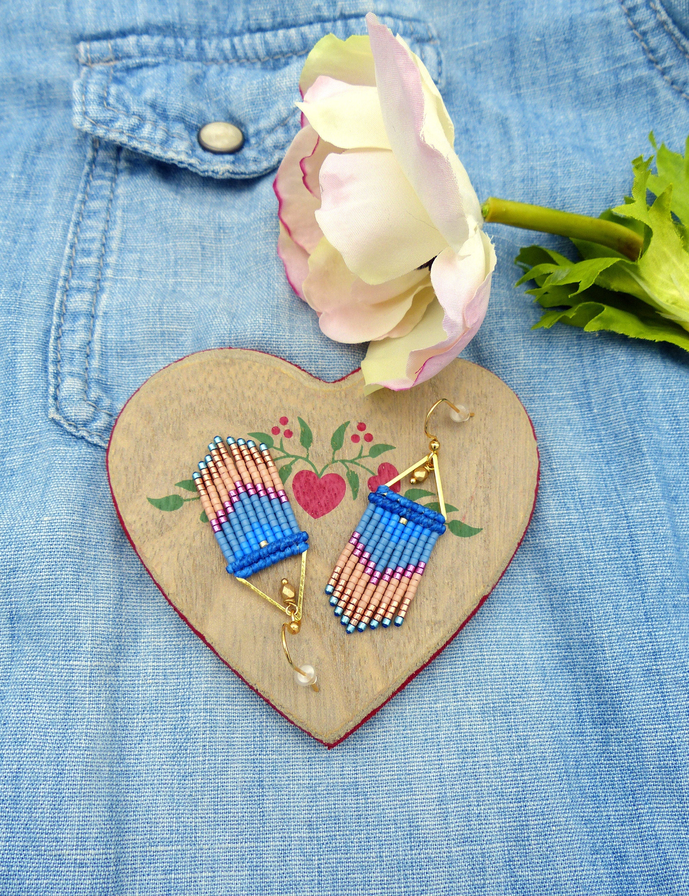 Peut inclure: Une paire de boucles d'oreilles faites à la main avec un design géométrique. Les boucles d'oreilles sont faites de perles bleues, roses et dorées et ont une forme triangulaire. Les boucles d'oreilles sont présentées sur une surface en bois en forme de cœur.