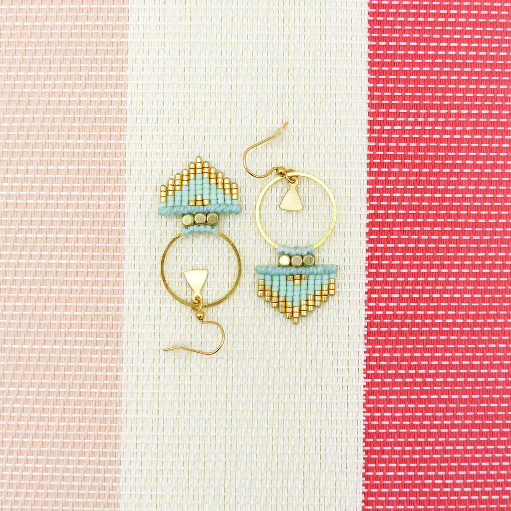 Peut inclure: Une paire de boucles d'oreilles créoles dorées avec un motif géométrique composé de perles bleu clair et dorées. Les boucles d'oreilles sont suspendues sur un fond rayé blanc et rose.