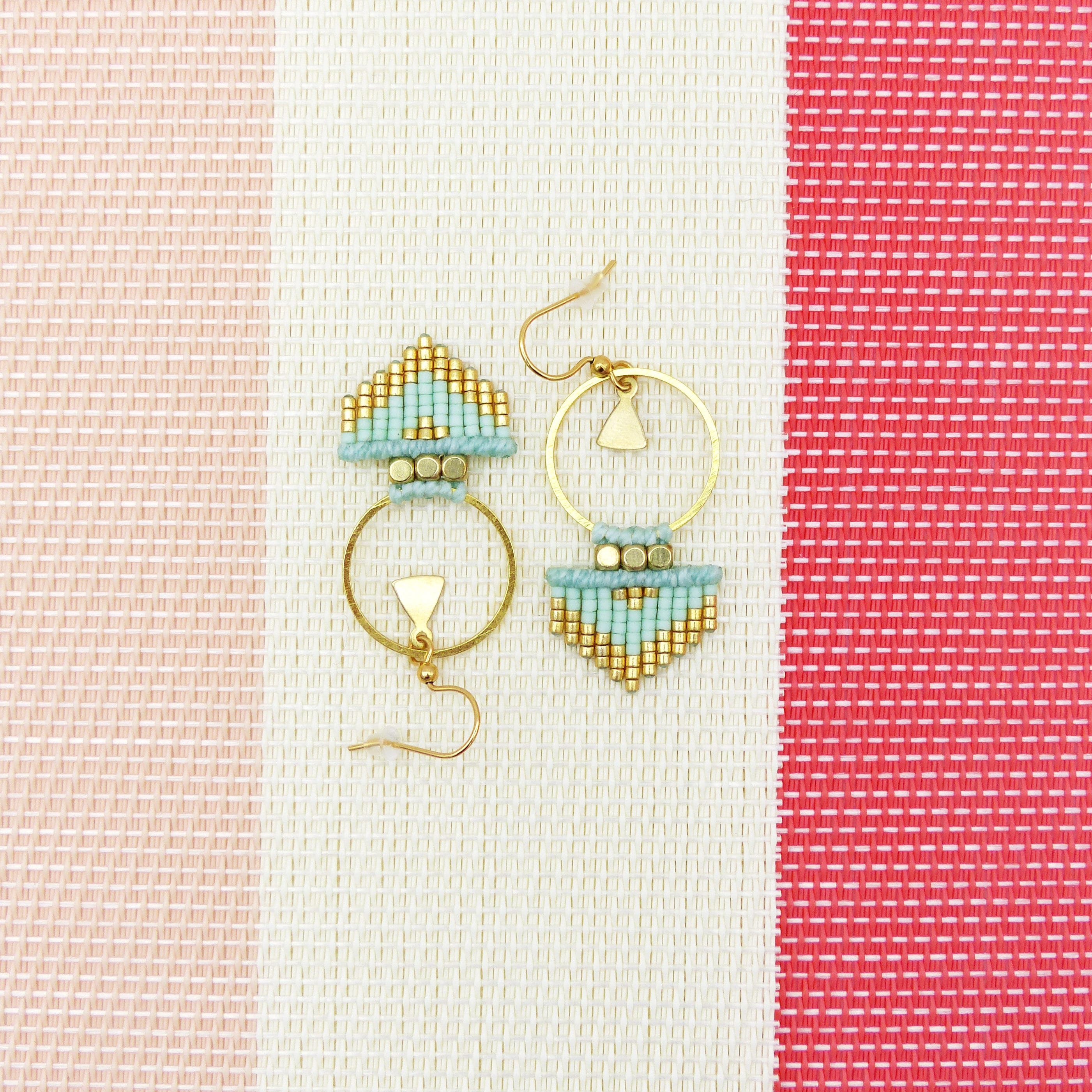 Peut inclure: Une paire de boucles d'oreilles créoles dorées avec un motif géométrique composé de perles bleu clair et dorées. Les boucles d'oreilles sont suspendues sur un fond rayé blanc et rose.