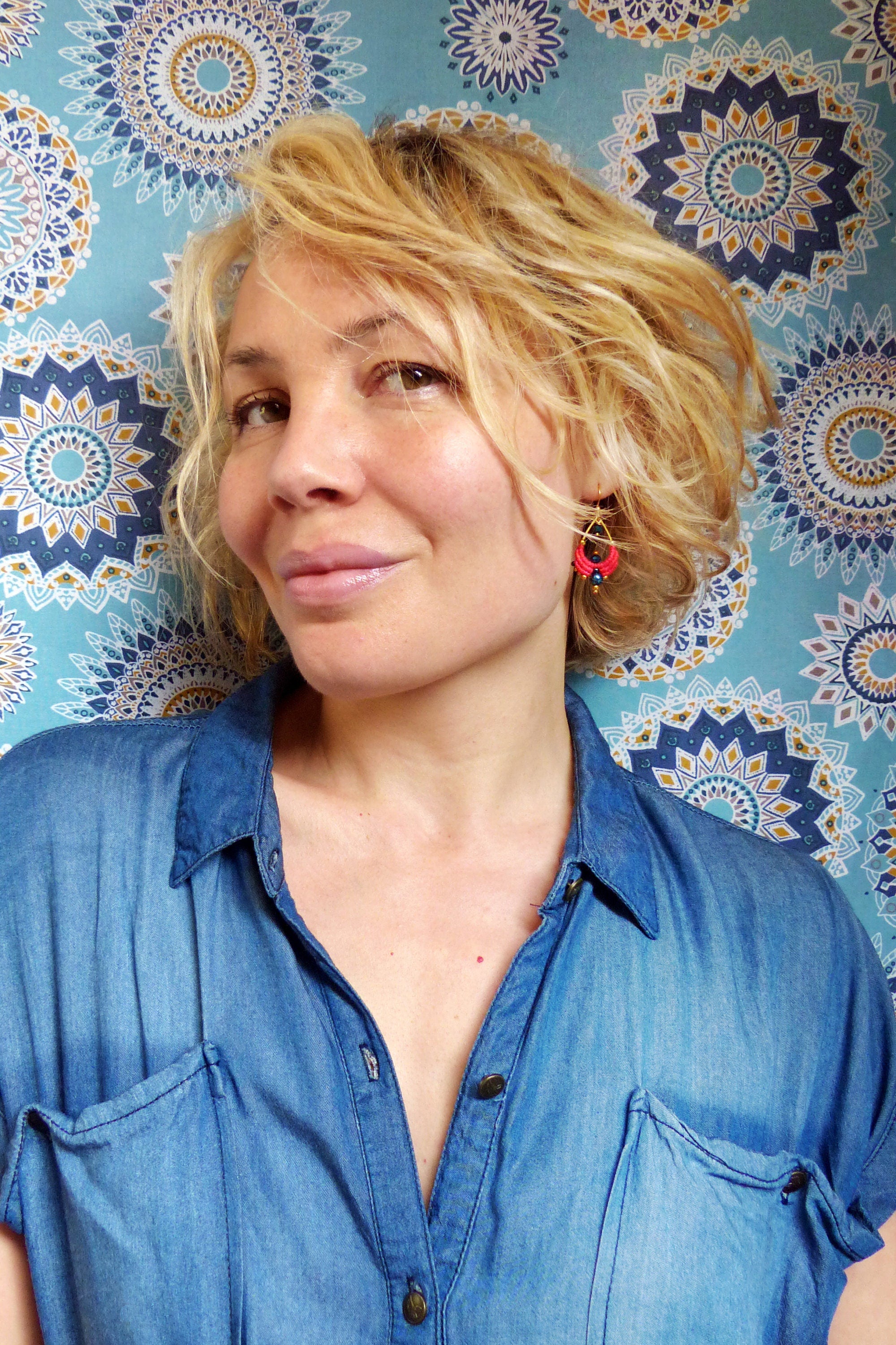Peut inclure: Une femme aux cheveux blonds portant une chemise en jean bleu et des boucles d'oreilles rouges. L'arrière-plan est un tissu à motifs bleus et dorés.