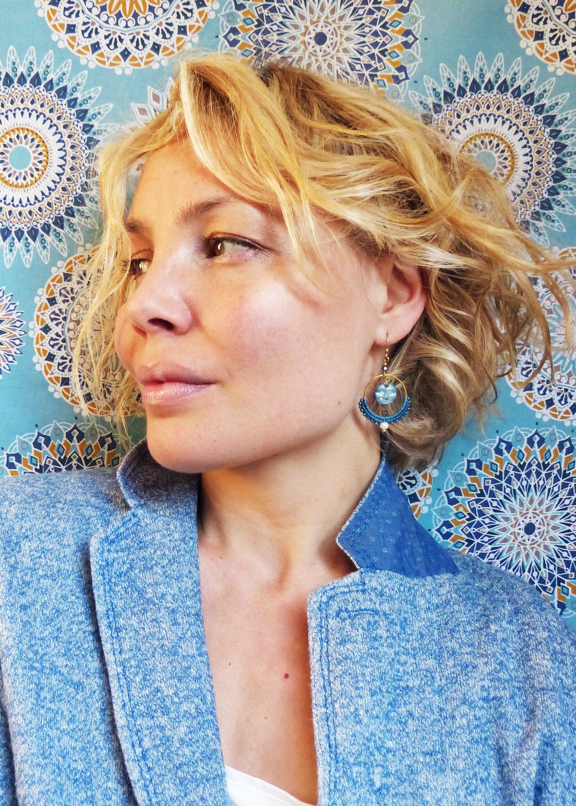 Peut inclure: Une femme aux cheveux blonds portant une veste en jean bleue et des boucles d'oreilles en perles bleues et dorées. L'arrière-plan est un tissu à motifs bleus et dorés.