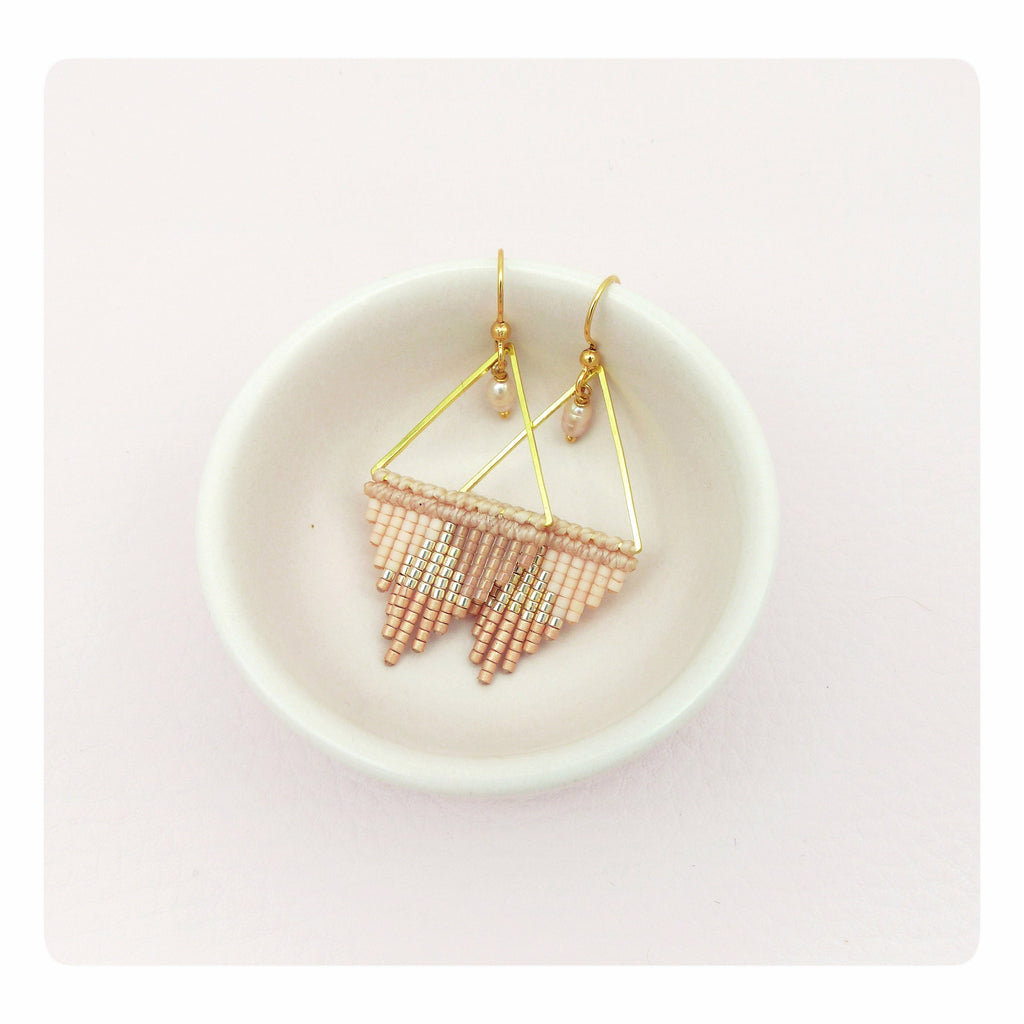 Peut inclure: Une paire de boucles d'oreilles triangulaires dorées avec une frange de perles roses et dorées. Les boucles d'oreilles sont présentées sur un plat en céramique blanche.