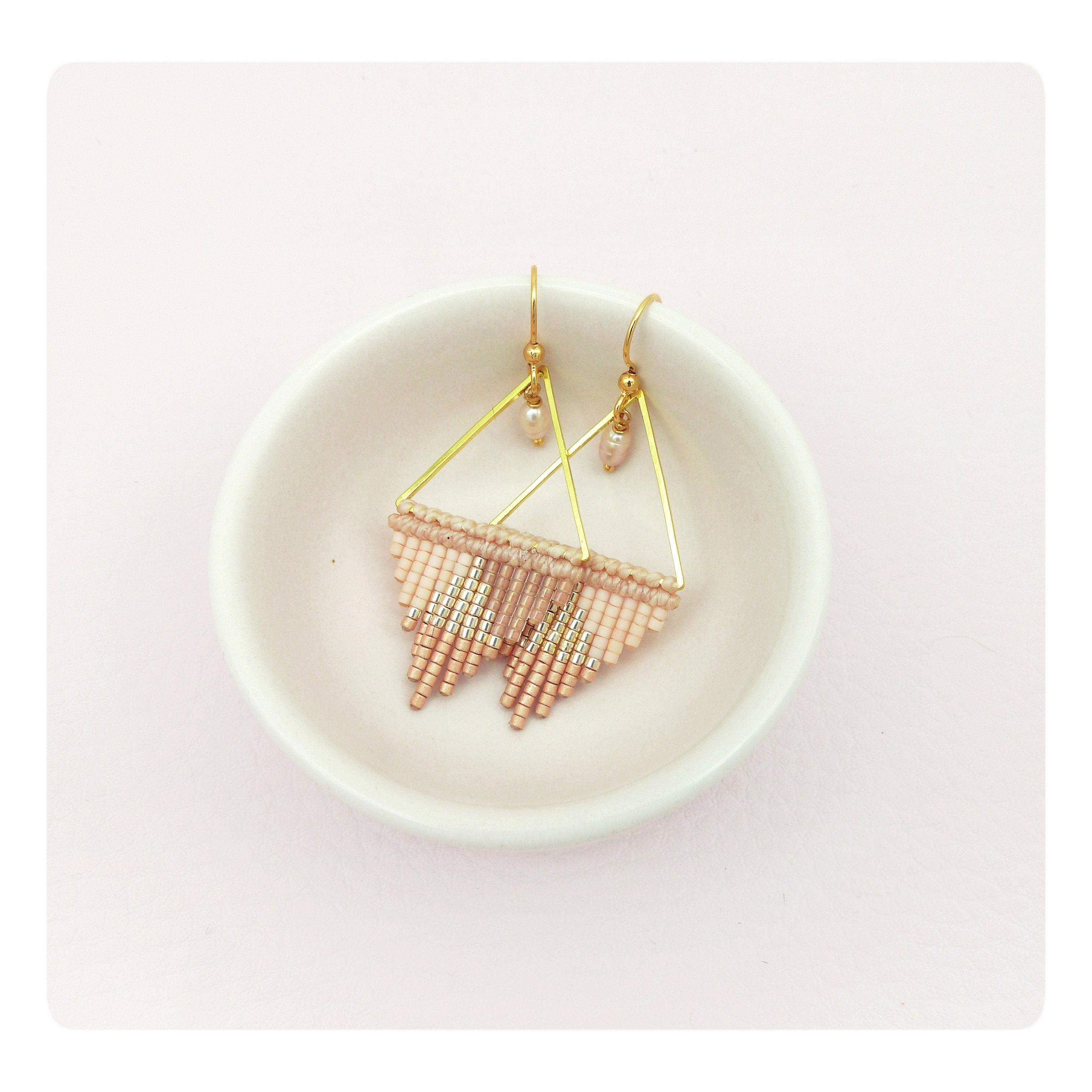 Peut inclure: Une paire de boucles d'oreilles triangulaires dorées avec une frange de perles roses et dorées. Les boucles d'oreilles sont présentées sur un plat en céramique blanche.
