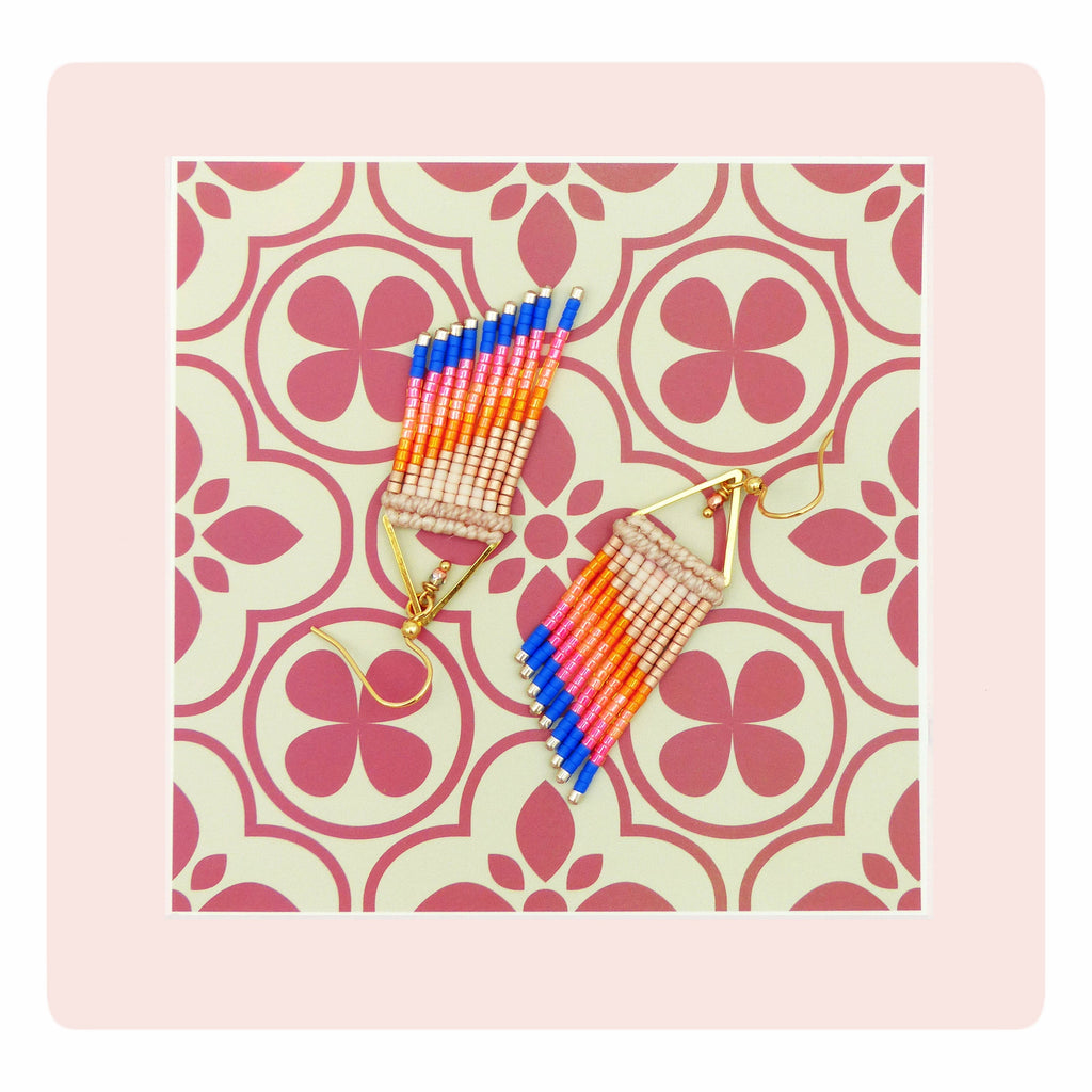 Peut inclure: Une paire de boucles d'oreilles à franges colorées. Les boucles d'oreilles sont composées de perles bleues, roses, oranges et beiges. Elles sont dotées de crochets d'oreilles dorés et sont présentées sur un fond à motifs.