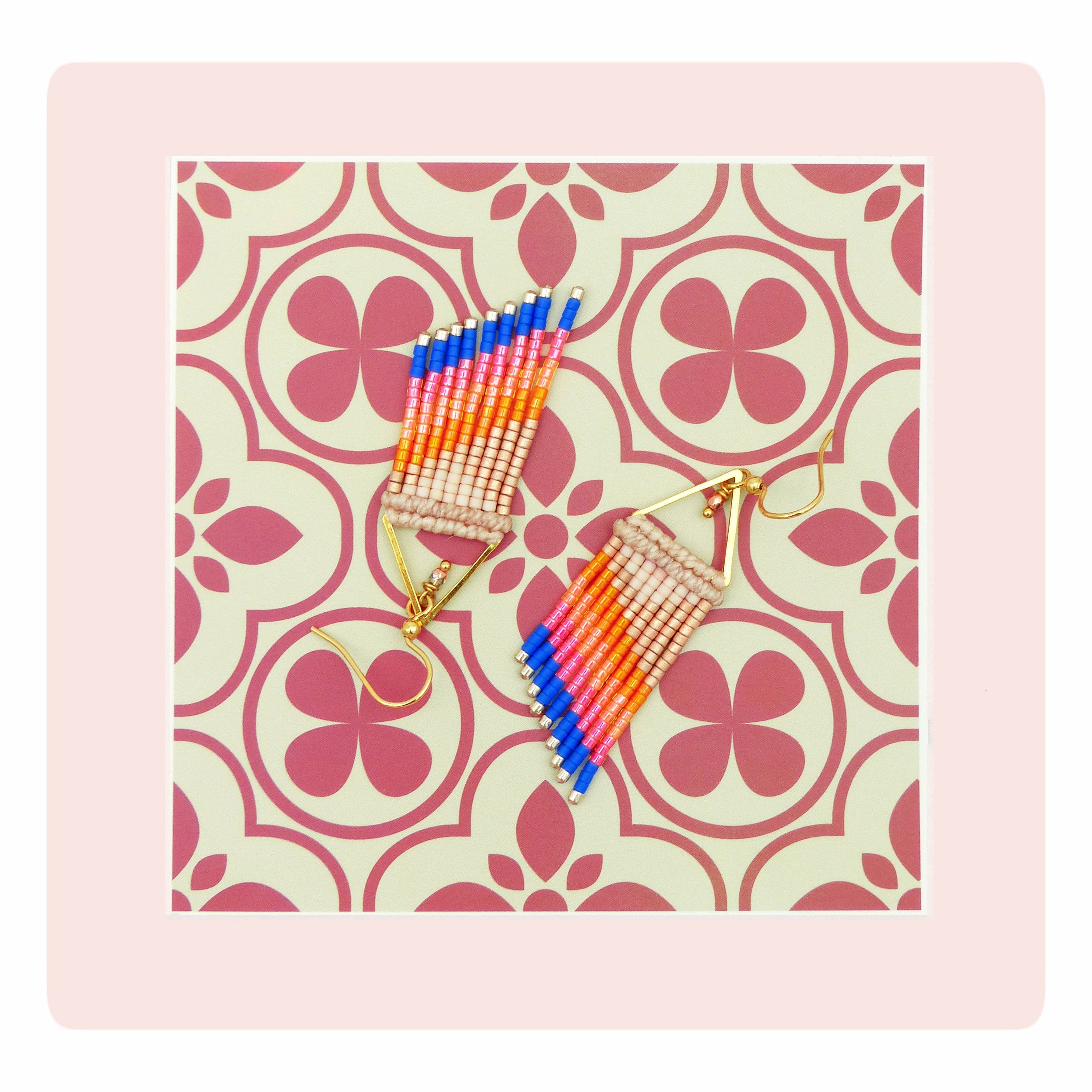 Peut inclure: Une paire de boucles d'oreilles à franges colorées. Les boucles d'oreilles sont composées de perles bleues, roses, oranges et beiges. Elles sont dotées de crochets d'oreilles dorés et sont présentées sur un fond à motifs.
