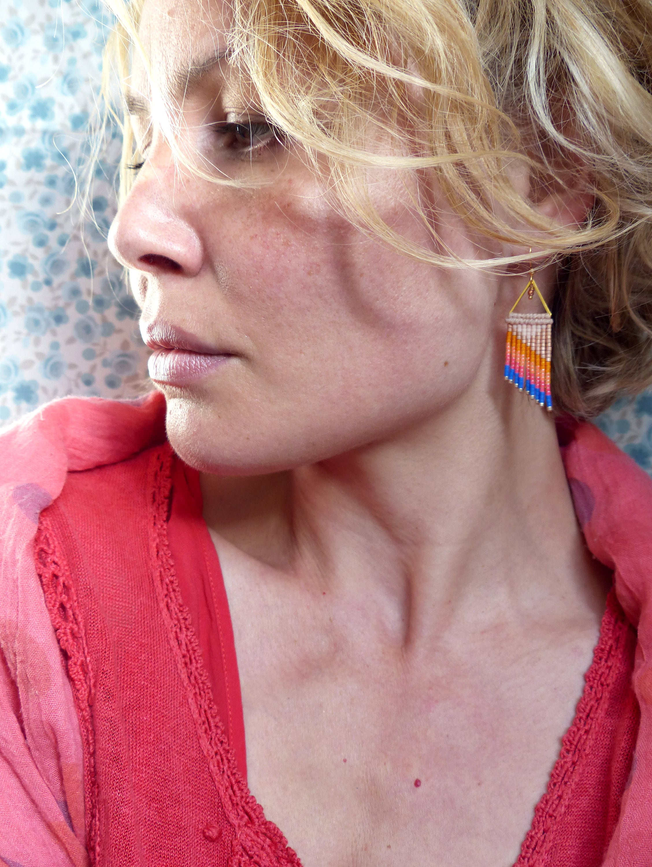 Peut inclure: Gros plan d'une femme portant des boucles d'oreilles en perles. Les boucles d'oreilles triangulaires sont dorées en haut et composées de perles roses, oranges, bleues et blanches. La femme porte un haut rouge et une écharpe rose.