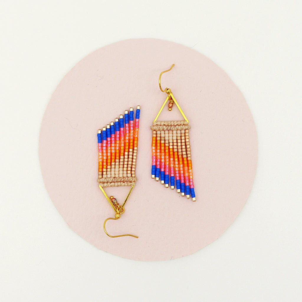 Peut inclure: Une paire de boucles d'oreilles perlées faites à la main. Les boucles d'oreilles présentent un motif géométrique avec une combinaison de perles bleues, oranges et beiges. Les boucles d'oreilles sont suspendues à des crochets et des cadres triangulaires dorés. Elles sont posées sur une surface rose.