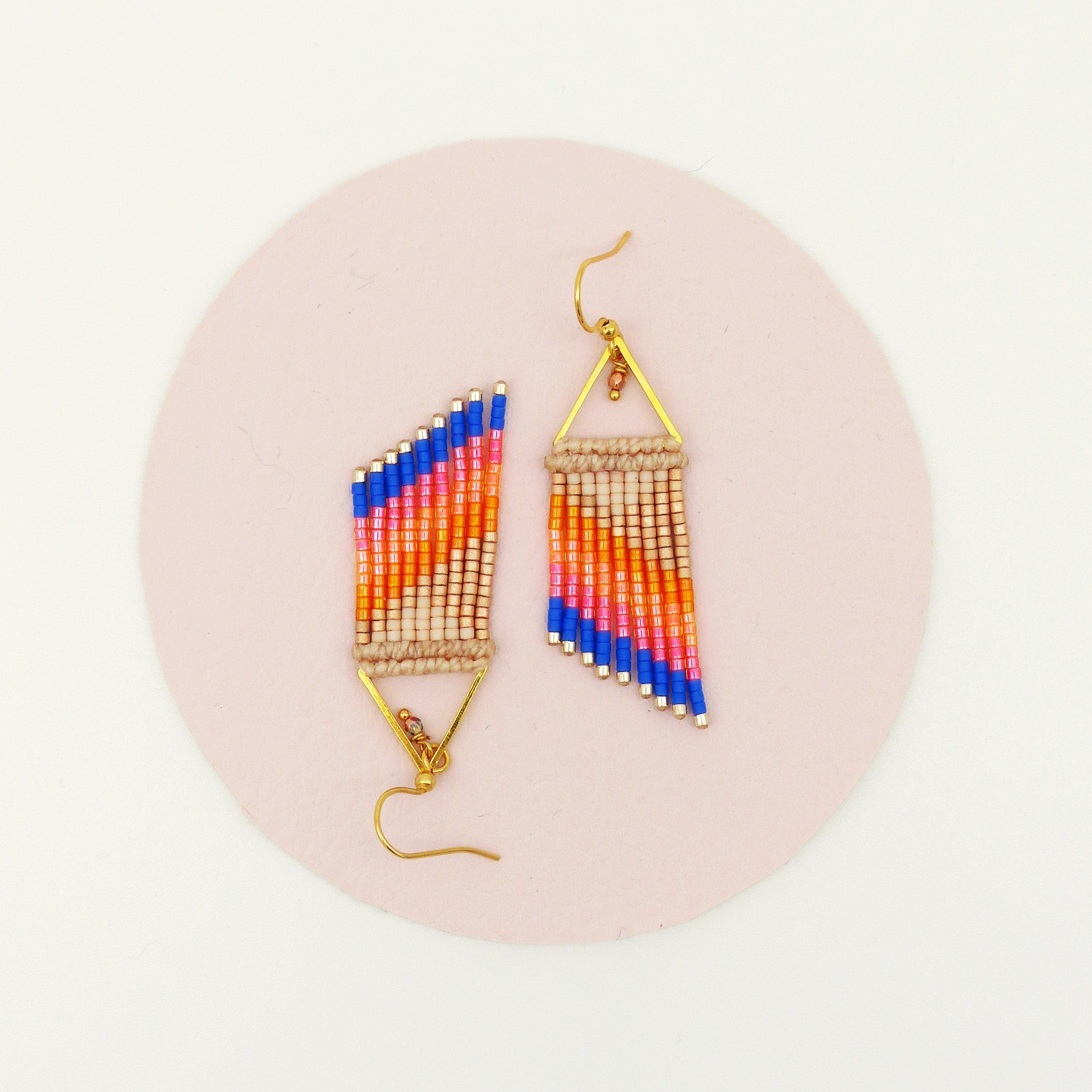 Peut inclure: Une paire de boucles d'oreilles perlées faites à la main. Les boucles d'oreilles présentent un motif géométrique avec une combinaison de perles bleues, oranges et beiges. Les boucles d'oreilles sont suspendues à des crochets et des cadres triangulaires dorés. Elles sont posées sur une surface rose.