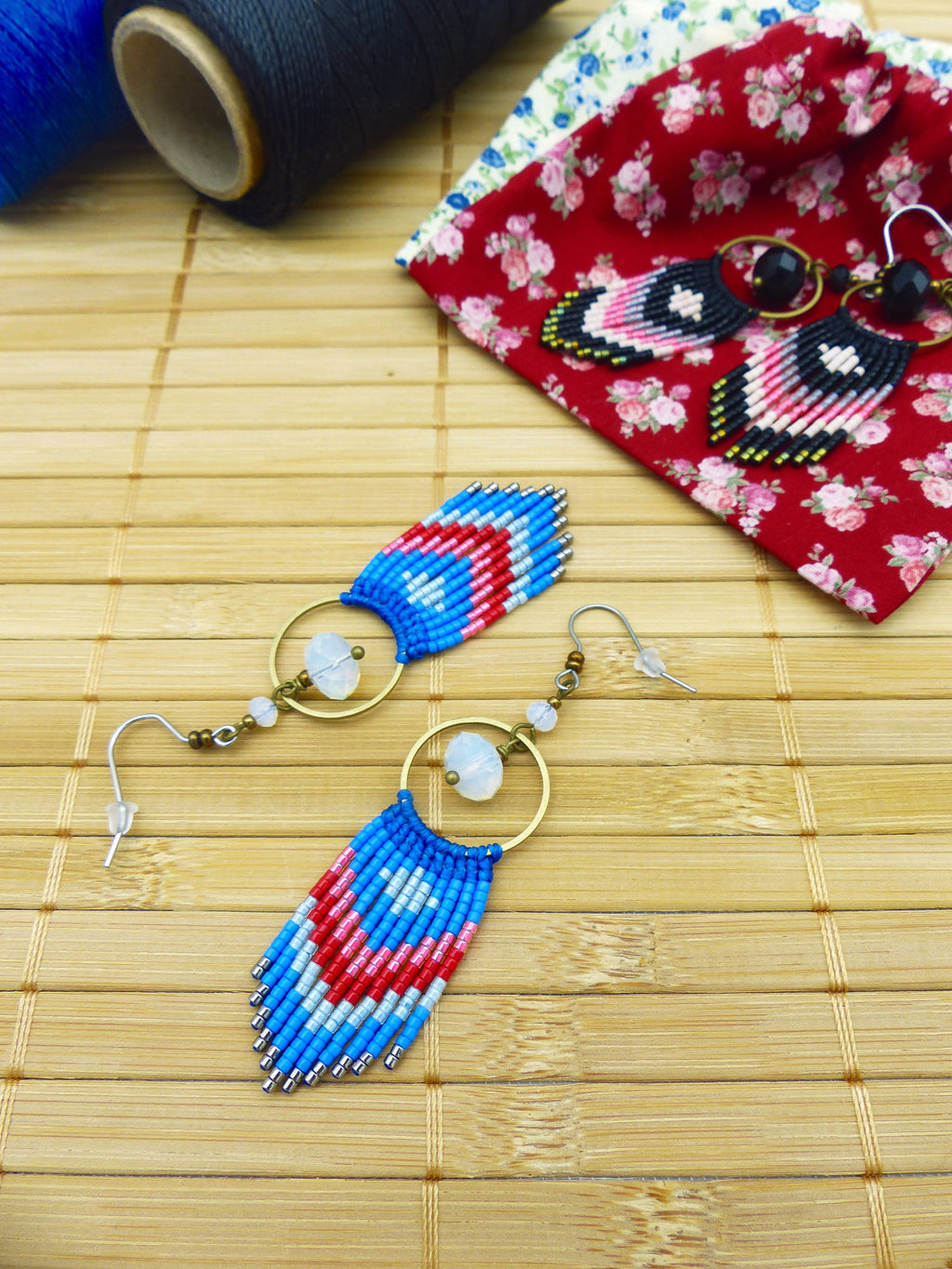 Peut inclure: Une paire de boucles d'oreilles en perles faites à la main avec un motif bleu, rouge et blanc. Les boucles d'oreilles ont une forme de larme avec une petite pierre ronde blanche au sommet. Les boucles d'oreilles sont suspendues à un anneau doré.