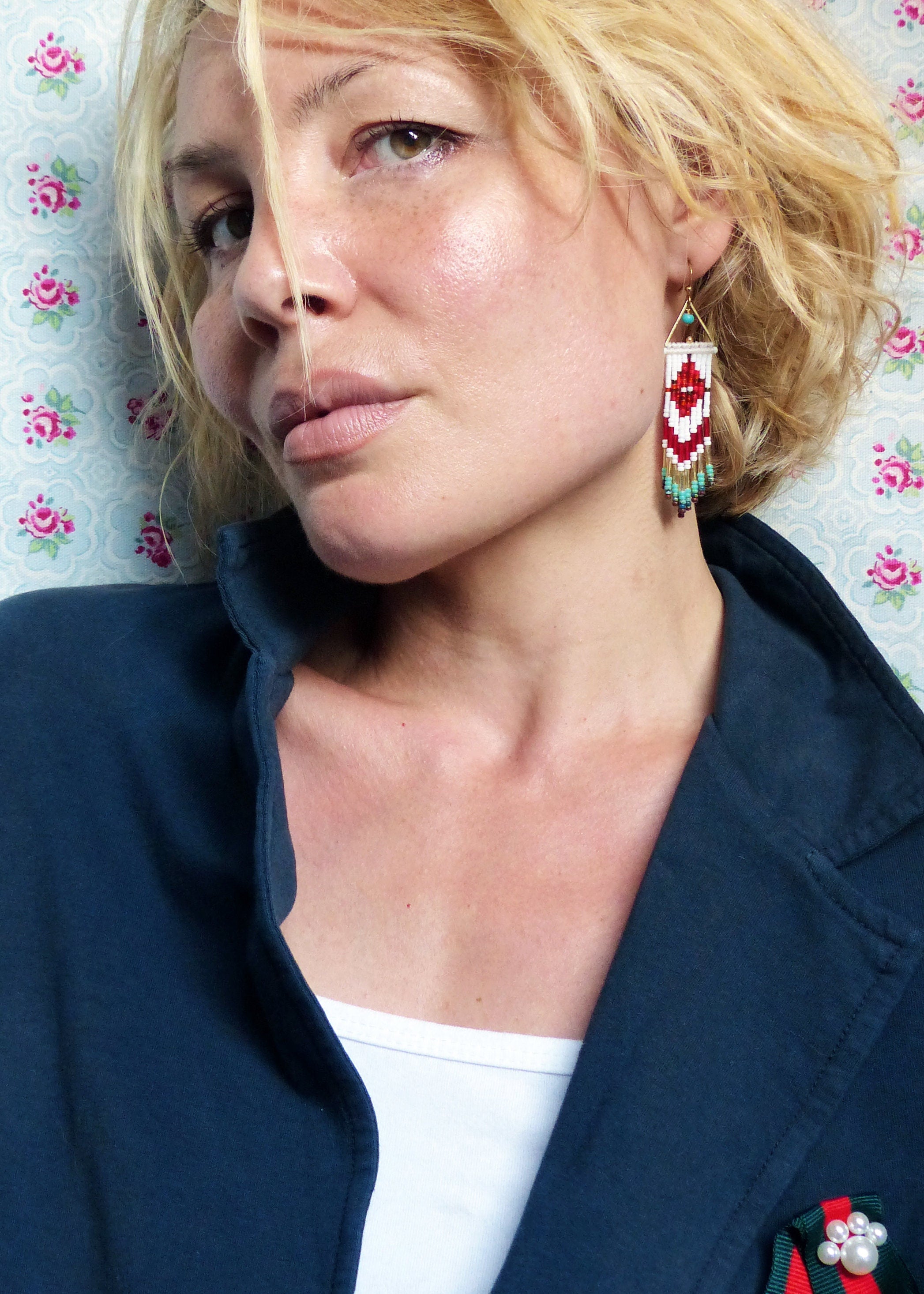 Peut inclure: Une femme aux cheveux blonds portant un blazer bleu foncé et un haut blanc. Elle porte de longues boucles d'oreilles en perles rouges, blanches et turquoise.