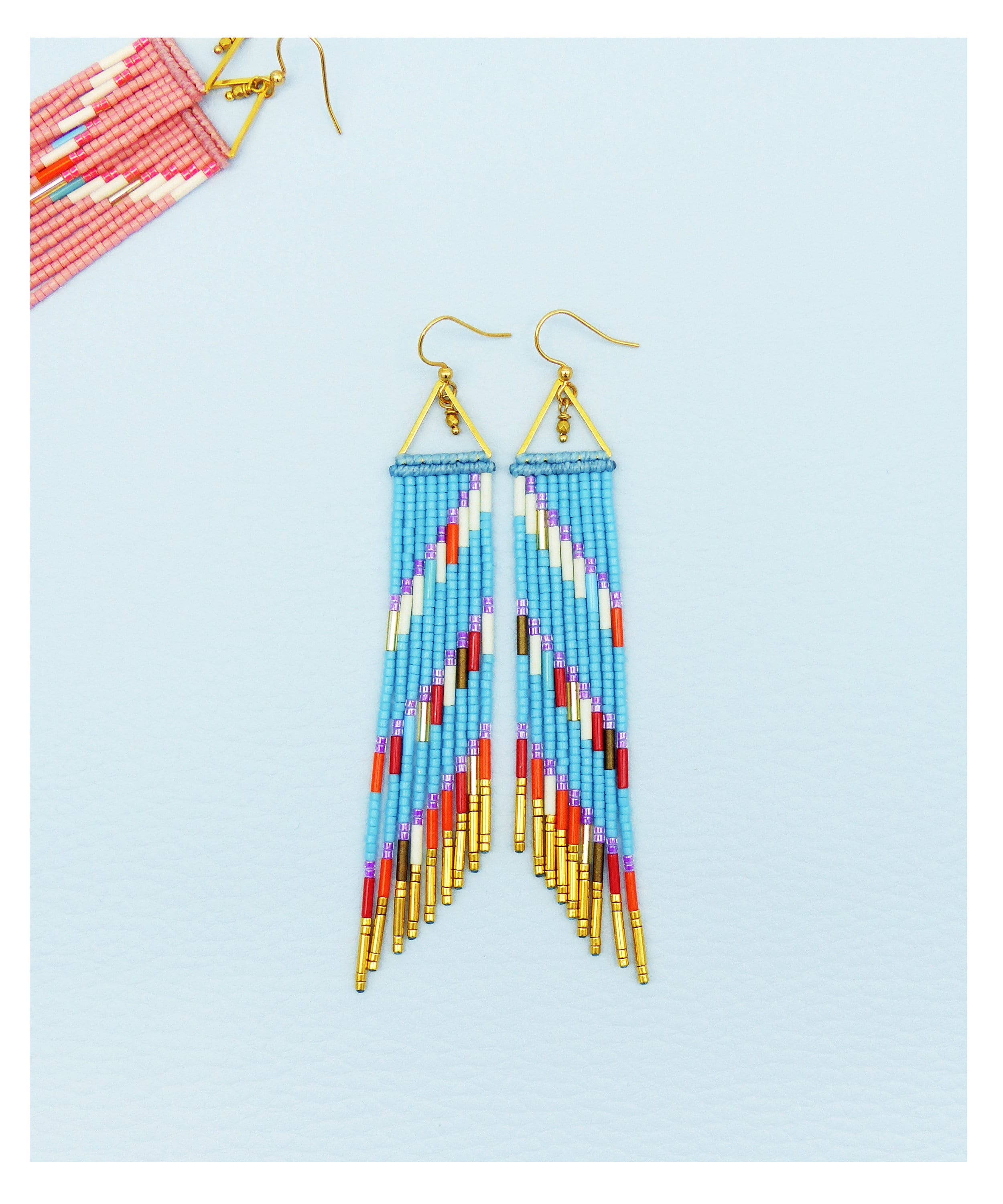 Peut inclure: Une paire de longues boucles d'oreilles en perles avec un motif géométrique. Les boucles d'oreilles sont faites de perles bleues, blanches, orange, rouges et dorées. Les boucles d'oreilles sont suspendues à des anneaux dorés en forme de triangle.