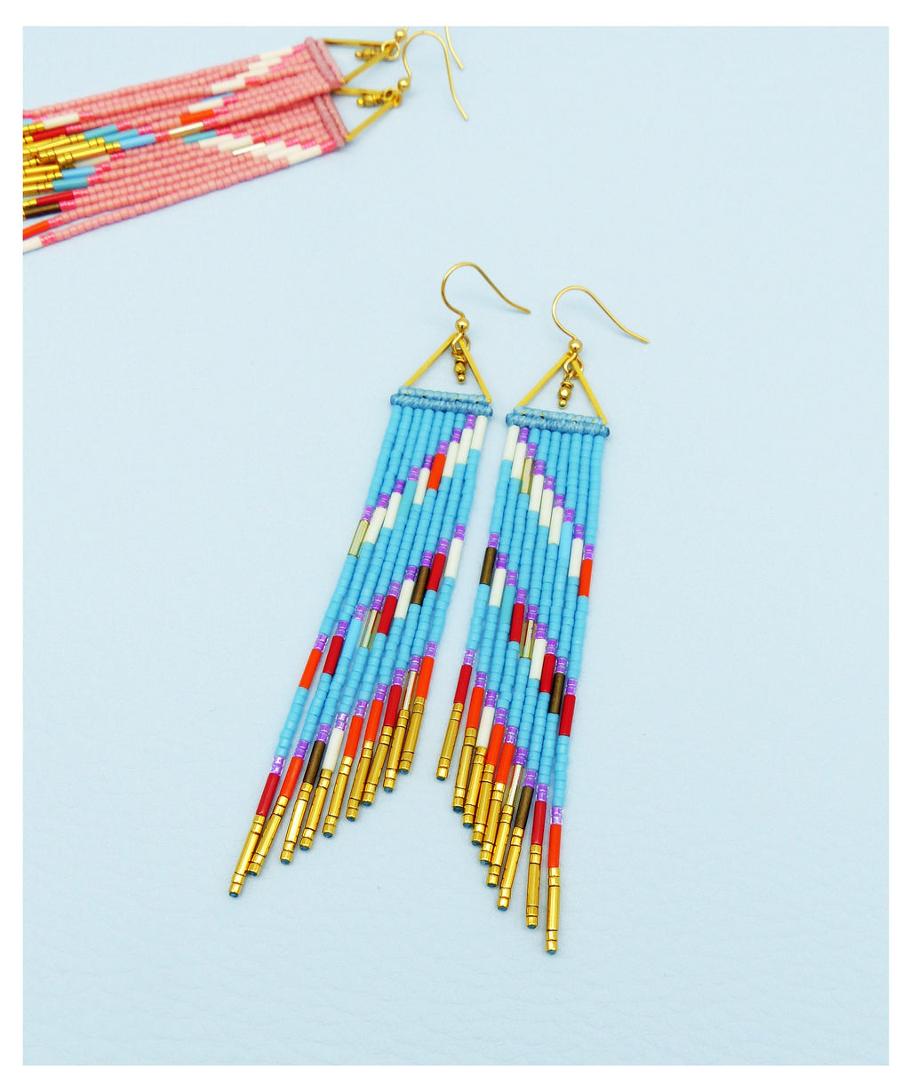 Peut inclure: Une paire de longues boucles d'oreilles en perles avec un motif géométrique. Les boucles d'oreilles sont faites de perles bleues, blanches, rouges, orange et violettes. Les boucles d'oreilles ont des accents dorés.